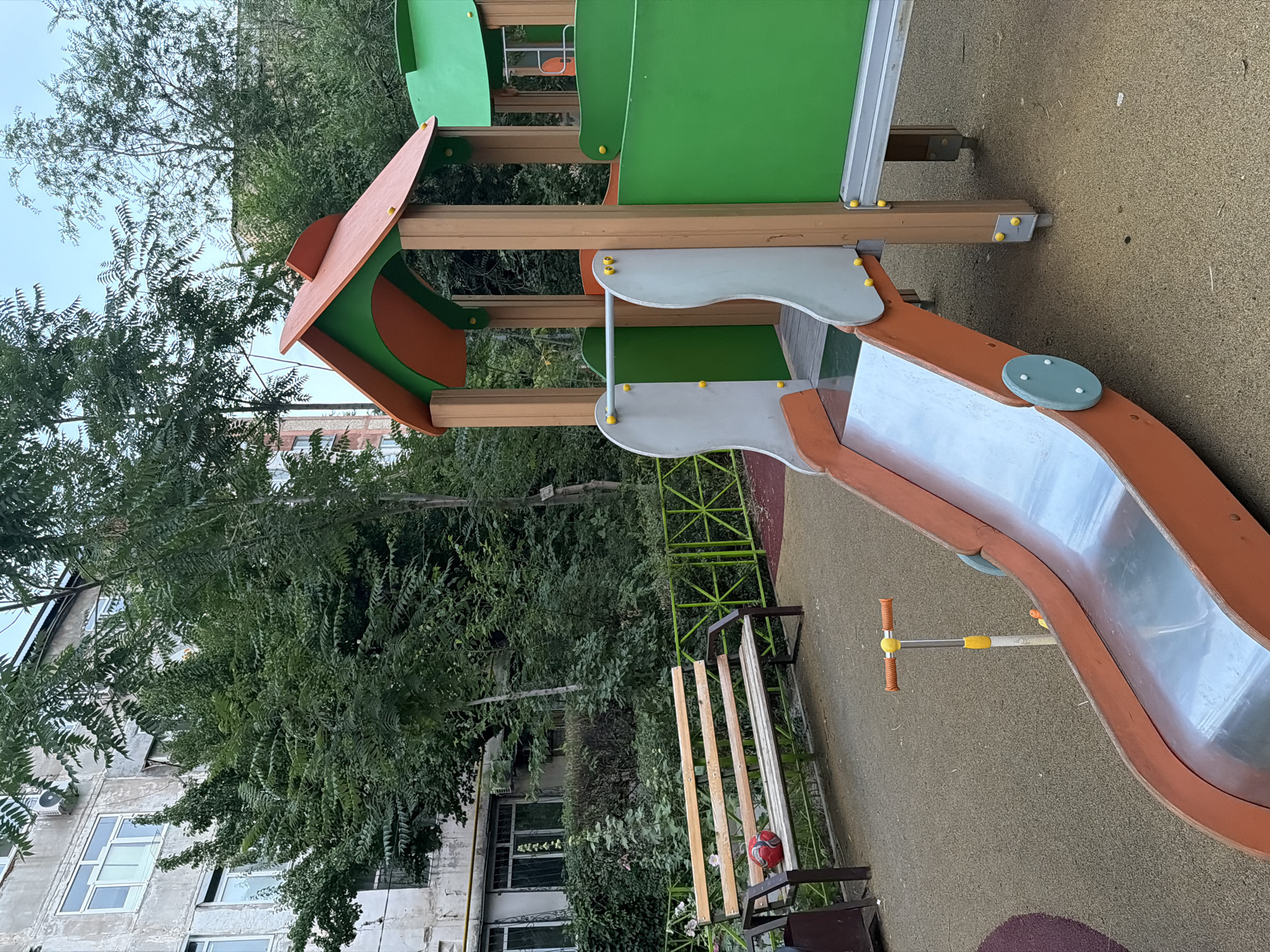 The image depicts a children's playground in Shymkent, Kazakhstan, situated in the vicinity of an apartment building. In the foreground, a metal and plastic slide with an orange frame is visible, positioned on a soft, textured ground surface. To the left of the slide, a scooter with orange handlebars and yellow accents stands upright. Further left, a wooden bench with metal legs holds a red ball. A green metal fence encloses the playground area. Behind the playground, dense green trees and bushes obscure the lower floors of a multi-story apartment building with a grey facade and white-framed windows. The sky is visible in the upper right corner, indicating an outdoor setting. The overall scene suggests a public recreational space within an urban residential area.