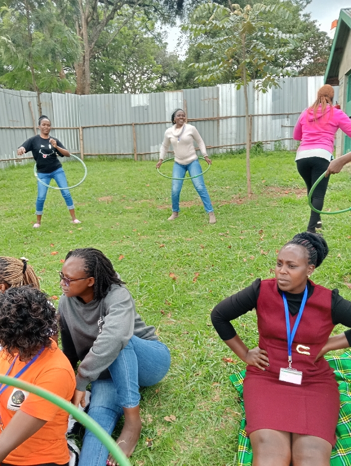 This image captures a group of women engaged in a casual outdoor activity on a grassy area in Riara Ridge, Kenya. The scene appears to be set during the daytime, under soft, possibly overcast, lighting.
In the foreground, two women are seated on the lush green grass. On the left, a woman with braided hair, wearing a grey long-sleeved sweatshirt and blue jeans, sits with her legs crossed and bare feet visible, looking towards the left. To her right, another woman in a maroon sleeveless dress layered over a black long-sleeved top, with a blue lanyard and white ID badge around her neck, is seated on a green, yellow, and black plaid blanket. She has her hands on her hips and is looking towards the right side of the frame. A third woman, wearing an orange t-shirt, is partially visible on the far left.
In the middle ground and background, three women are actively participating in hula-hooping. On the left, a woman in a black t-shirt featuring a white graphic (possibly a smiling face) and blue jeans is smiling broadly while swinging a green hula hoop around her waist. To her right, another woman, also smiling, is hula-hooping while wearing a light-colored (possibly beige) long-sleeved top and blue jeans. On the far right, partially visible, a woman in a bright pink hoodie and dark pants is also engaged in hula-hooping.
The background is defined by a corrugated metal fence, behind which several trees are visible. A small building or structure is partially seen on the far right. The overall atmosphere of the image suggests a relaxed and social gathering, with participants enjoying recreational activities outdoors. Visible text on the ID badges and clothing is not clearly legible.
