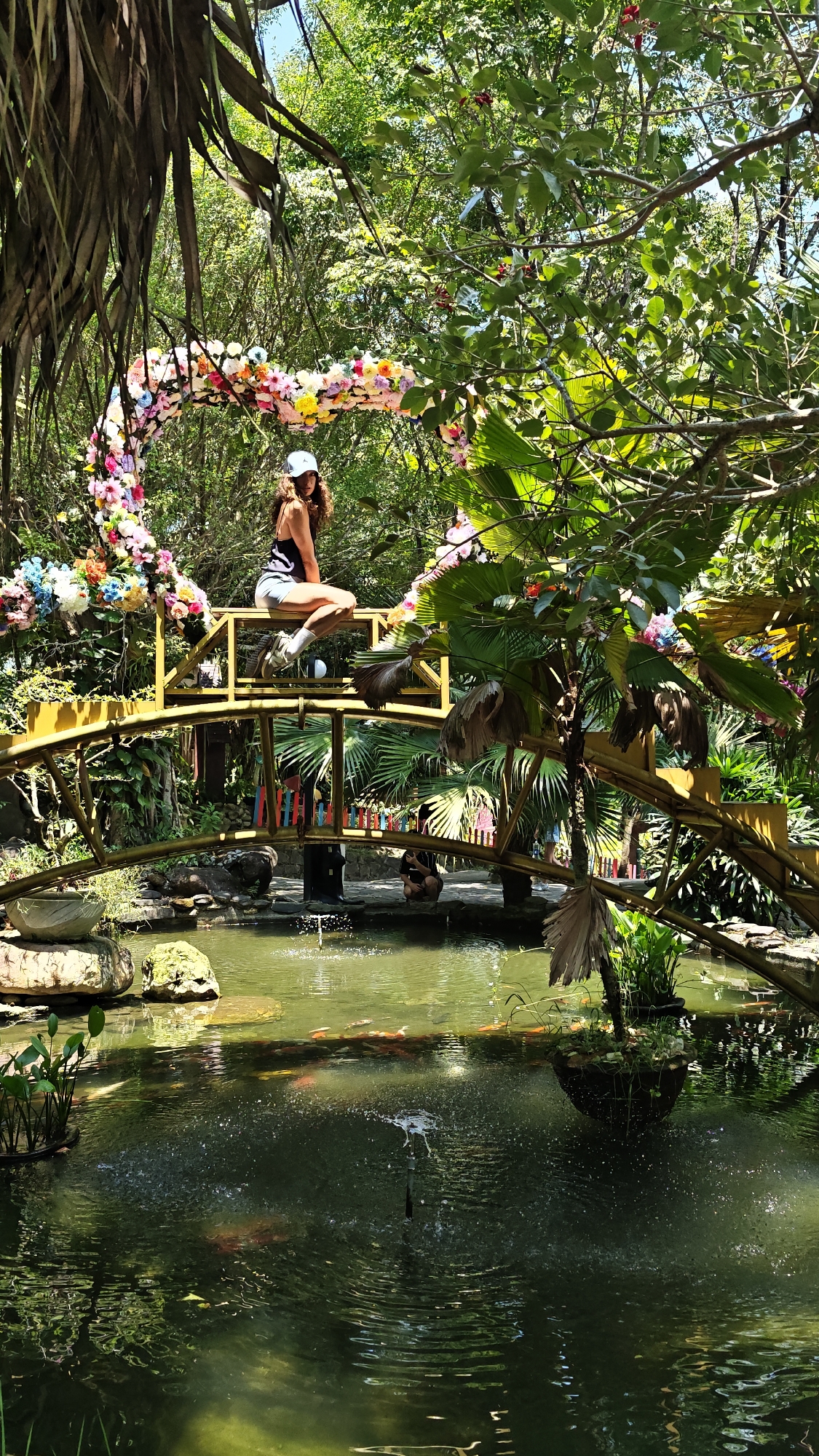 A woman sits on a yellow bridge in the Ba Na Hills of Đà Nẵng, Vietnam. The bridge is decorated with a heart-shaped floral archway. Below the bridge is a pond with a fountain and koi fish. Green trees and plants surround the area.