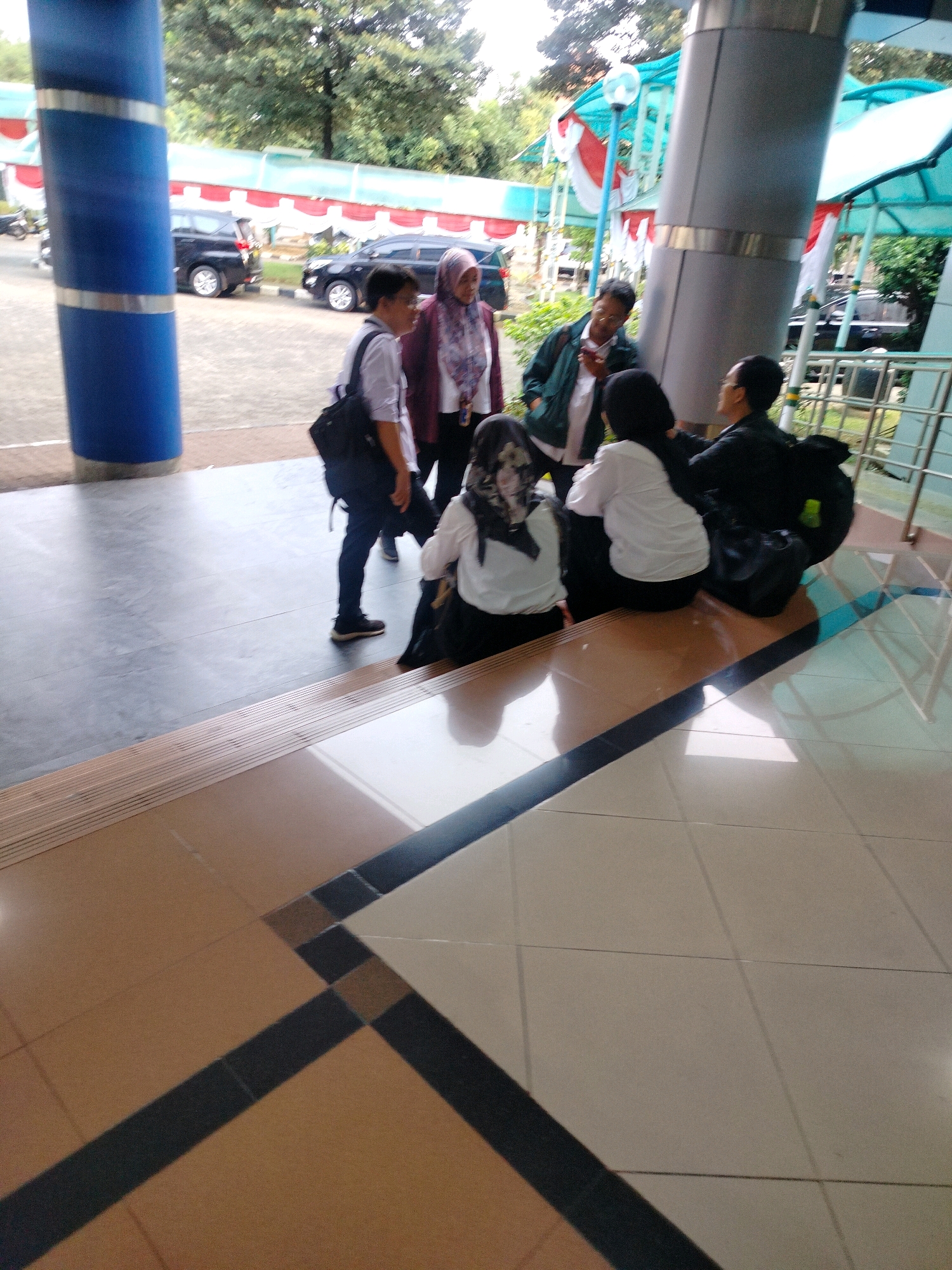 The image depicts a group of five individuals gathered near a building entrance, likely in East Jakarta, Indonesia, given the context. Two individuals are standing and facing the other three who are seated on steps. One standing person, on the left, wears a light-colored shirt and dark pants, carrying a black backpack. Another standing person, in the center, is wearing a green jacket over a white shirt and glasses. The three seated individuals are facing away from the camera. The person on the left is wearing a light-colored shirt and a headscarf. The middle seated person wears a white shirt and dark pants, with a patterned headscarf. The person on the right is dressed in dark clothing. The environment appears to be outdoors, with trees and buildings visible in the background. The foreground consists of tiled flooring and steps. A blue cylindrical pillar is visible on the far left. Several cars are parked in the background. The overall scene suggests a casual gathering or a waiting area.