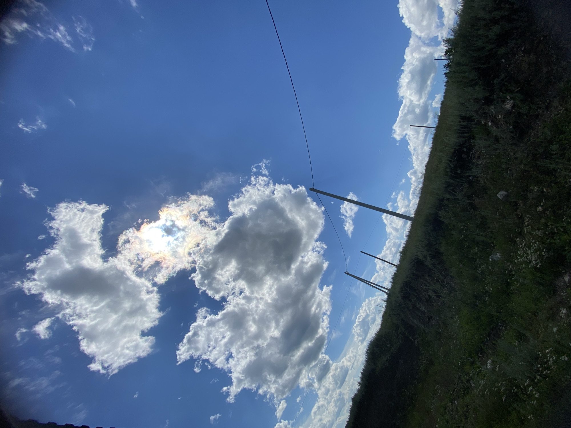 A low-angle, wide shot captures a bright, partly cloudy sky above a grassy hillside. The sun is visible, partially obscured by a large, fluffy cloud, casting a halo of light that refracts into subtle rainbow colors. A thin, dark wire stretches horizontally across the upper portion of the frame, supported by utility poles. The hillside is covered in dense, green vegetation, with some scattered white wildflowers. The overall scene suggests an outdoor, possibly rural setting in the city of Yuldybayevo, Russia.