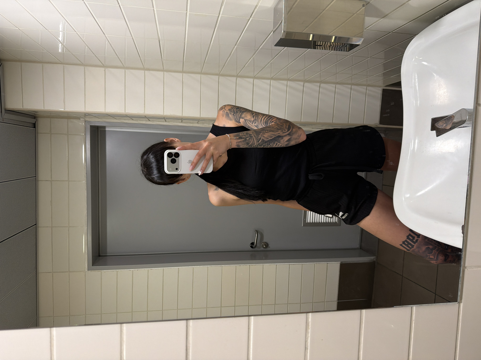 The image depicts a mirror selfie taken by a person in a public restroom located in Plovdiv, Bulgaria. The individual, whose face is obscured by a white smartphone held in their left hand, is reflected from the waist up. They have dark hair, visible in a ponytail, and wear a black ribbed tank top and black shorts with a visible drawstring. Distinctive features include a full sleeve tattoo on the left arm, a smaller tattoo on the right shoulder, a dark tattoo in gothic script on the right lower leg, partially legible as "LIBERTAD," and another dark tattoo on the back of the left hand. The left wrist sports a thin silver bracelet with a charm, and the fingernails are manicured with white tips.
The background consists of walls tiled with light beige or off-white rectangular ceramic tiles, exhibiting horizontal and vertical grout lines. A light-grey door with a silver handle is visible behind the person, to the left, and features a dark rectangular vent near its bottom. The ceiling comprises grey, perforated panels, indicative of a suspended ceiling system. In the immediate foreground, a white ceramic sink with a silver faucet is partially visible, cut off by the bottom of the mirror. To the right of the person, a silver, wall-mounted dispenser is affixed to the tiled wall. The floor, as reflected in the mirror, appears to be covered with dark grey or brown tiles. The environment is uniformly lit by artificial lighting.