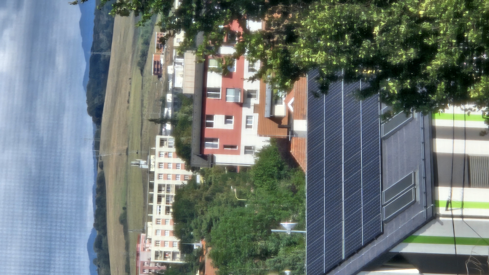This is a vertical, eye-level, medium shot of a residential area in Prešov, Slovakia. In the foreground, the dark, reflective surface of a rooftop covered with solar panels dominates the lower portion of the frame. Two skylights are visible on the grey rooftop just below the solar panels. A building with white and green vertical stripes is partially visible on the left side of the foreground. In the midground, several apartment buildings are nestled among lush green trees. One prominent building features a red and white facade with multiple windows. Further in the background, rolling hills with scattered trees stretch towards distant, hazy blue mountains under a cloudy sky. Power lines and transmission towers are visible on the hills. The overall impression is that of a suburban landscape.