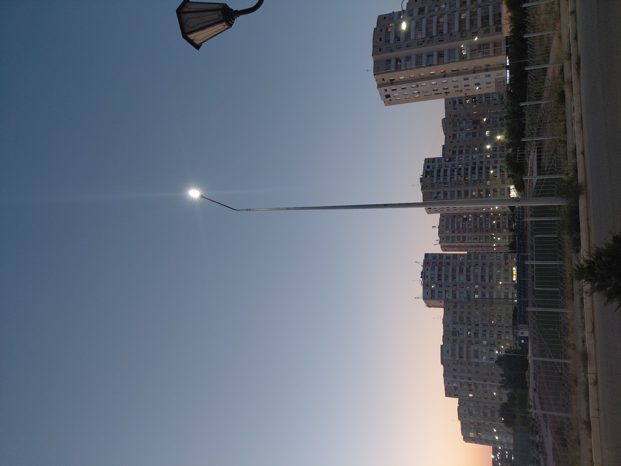 This is a vertical, wide shot taken at dusk in Aktau, Kazakhstan, showing a row of multi-story residential buildings. The sky is a gradient of deep blue at the top transitioning to a lighter, pale orange near the horizon. A tall, modern street lamp with a bright LED light stands prominently in the center-left of the frame, its light creating a lens flare. To the right, a vintage-style street lamp with a hexagonal glass enclosure is partially visible against the sky. In the foreground, there is a paved road and a sidewalk, separated by a low fence and some sparse vegetation. Behind the fence, there is a grassy area with trees and what appears to be a sports field with a net. The buildings are characteristic of Soviet-era architecture, with numerous windows and balconies. Some windows are illuminated, suggesting that people are inside. There are no people visible in the image. The scene depicts a typical urban residential area during twilight.