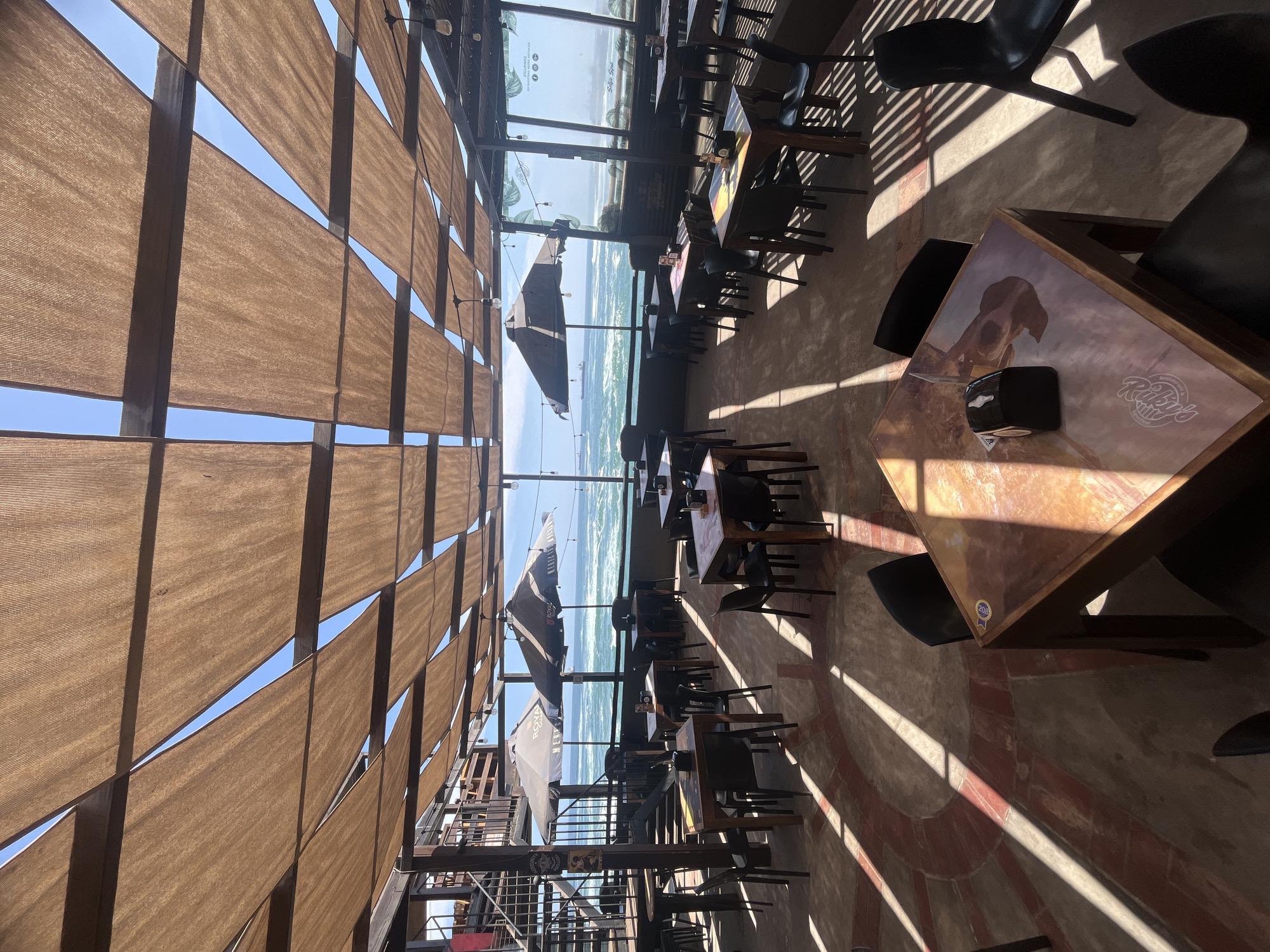 The image depicts the interior of a restaurant with a view of the ocean in Arica, Chile. The restaurant has a semi-outdoor structure with a slatted awning providing shade and glimpses of the sky. Numerous empty tables and chairs are arranged throughout the space, which is illuminated by natural light and shadows. The floor appears to be a combination of concrete and brick. Through a window, the ocean is visible. In the foreground, a square table with a restaurant logo is set with a napkin dispenser. The restaurant appears unoccupied at the time the photo was taken.
