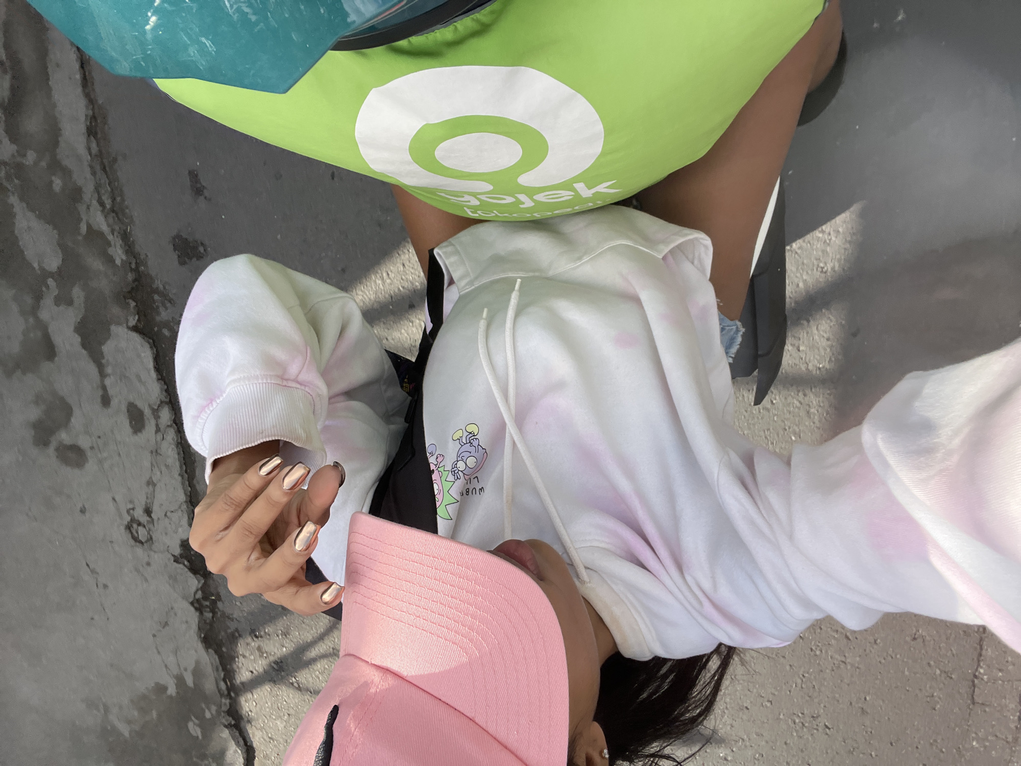 A person is seated on a vehicle, likely a motorcycle, in an outdoor setting. They are wearing a white tie-dye hoodie with pink accents, denim shorts, and a pink baseball cap. Their hands are visible, with metallic rose gold nail polish. In front of them is a green bag with a white circular logo, which reads "Gojek". The ground appears to be paved, suggesting an urban or semi-urban environment in North Kuta, Indonesia. The person is looking down, and their right arm is extended towards the camera, possibly taking a selfie. The overall lighting suggests it is daytime.