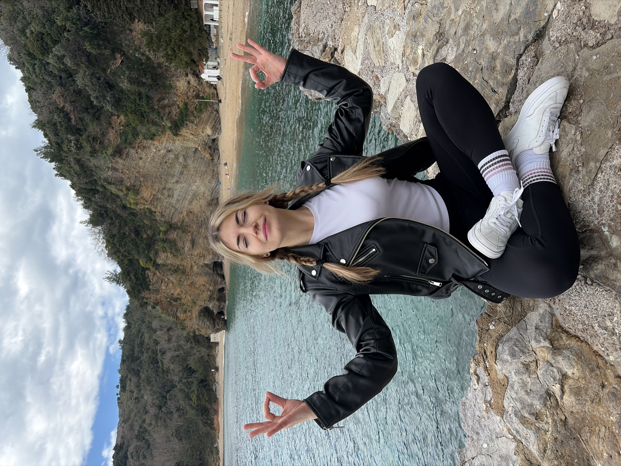 A young woman with blonde braided hair is sitting cross-legged on a rocky outcrop with the sea and a beach in the background. She is wearing a white T-shirt under a black leather jacket, black leggings, white socks with stripes, and white sneakers. She is making the "OK" hand gesture with both hands. The background shows a coastline with cliffs covered in greenery, a beach, and turquoise water. The location is likely near Budva, Montenegro, judging by the landscape. The weather appears overcast.