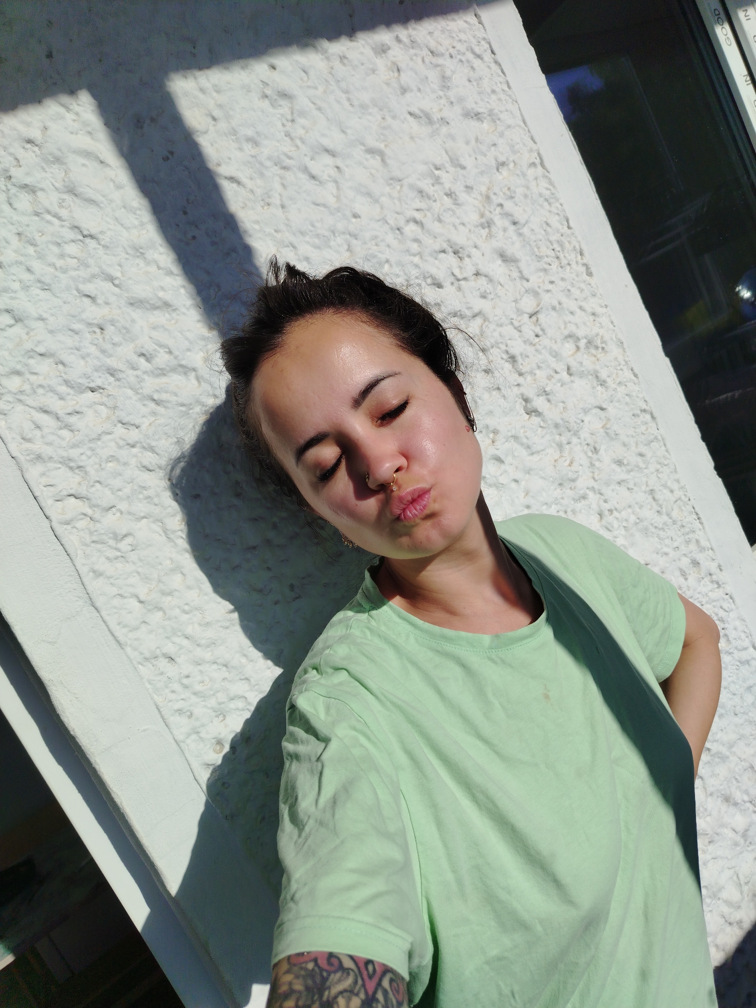 The image shows a young woman taking a selfie outdoors. She has dark hair pulled back into a bun and is wearing a light green t-shirt. Her eyes are closed, and her lips are pursed in a kiss. A nose ring is visible. She is leaning against a textured white wall, with a shadow cast across her. To her right, a window with dark panes is visible. The scene appears to be in the city of Noviy, Russia, based on the context provided, however there are no visual cues in the image to confirm this location.