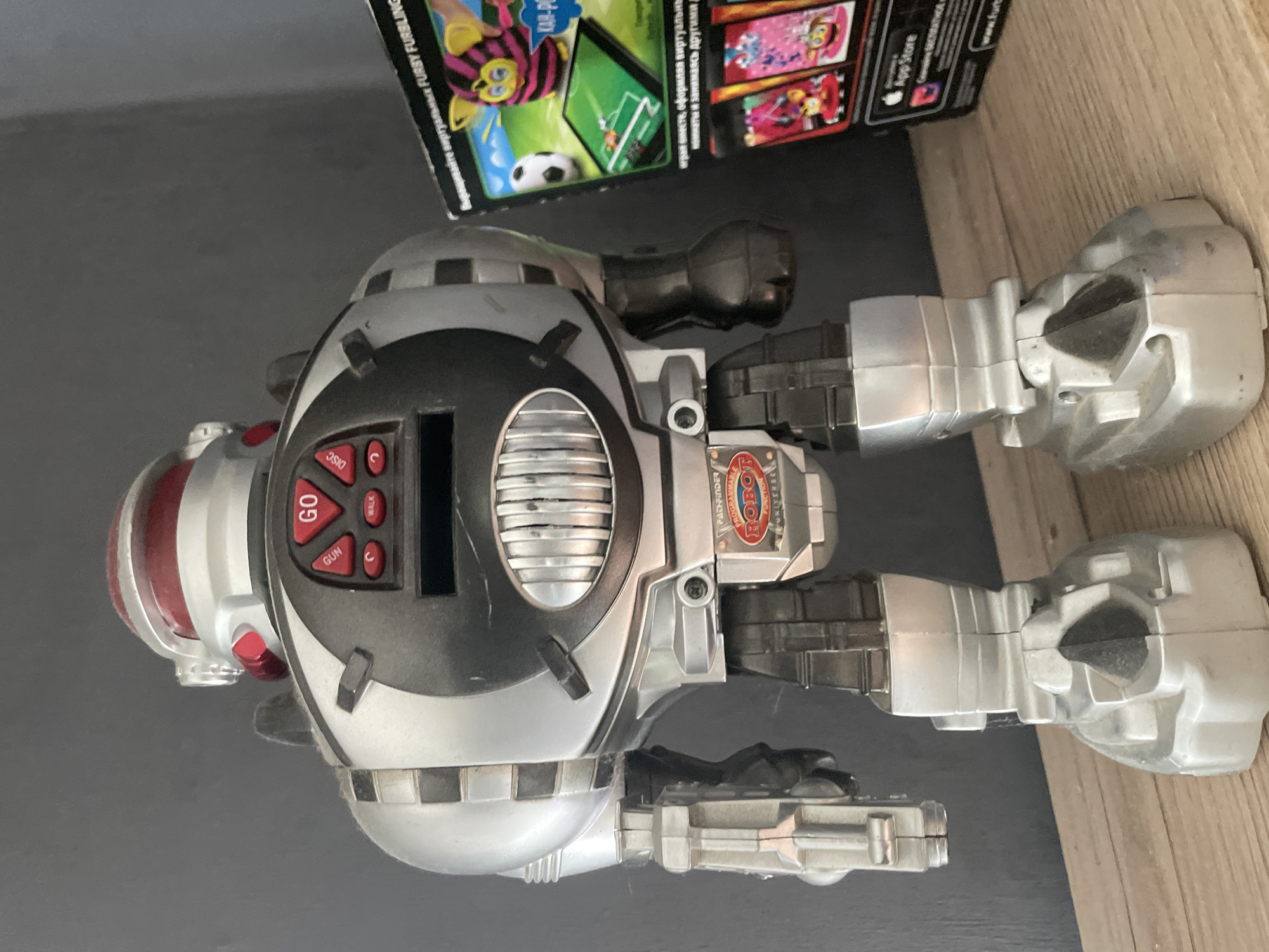A silver and red robot toy is standing on a wooden surface in front of a gray wall. The robot has a large, rounded body with a black panel on its chest featuring several red buttons and a small screen. It has two arms, one of which is extended forward, and two legs with blocky feet. To the right of the robot, a portion of a box with images of a Furby and a soccer game is visible. The box has text in Russian.