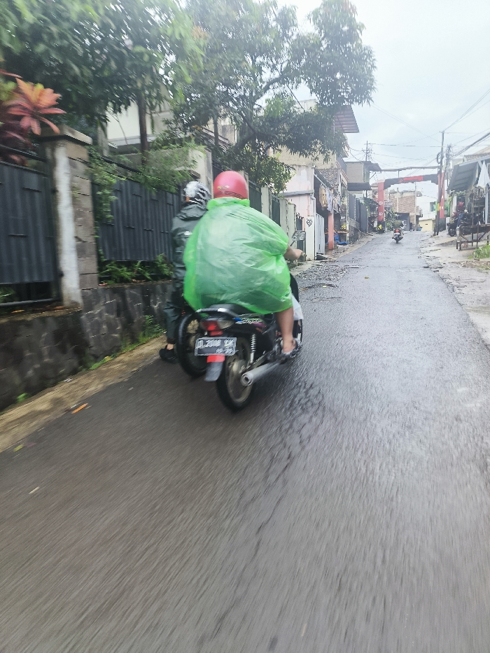 The image shows two people riding a motorcycle on a wet road in Cimenyan, Indonesia. The person driving the motorcycle is wearing a bright green raincoat and a red helmet. The other person riding on the back is wearing a dark jacket. Both people are not visible from the face due to the angle of the image. They are traveling along a narrow, paved road lined with buildings on either side. There are trees and vegetation in the background. The road is wet from recent rain, and the street is mostly empty. The motorcycle is moving forward at a moderate speed. The license plate of the motorcycle can be seen at the back of the image as DL 3040 BK.