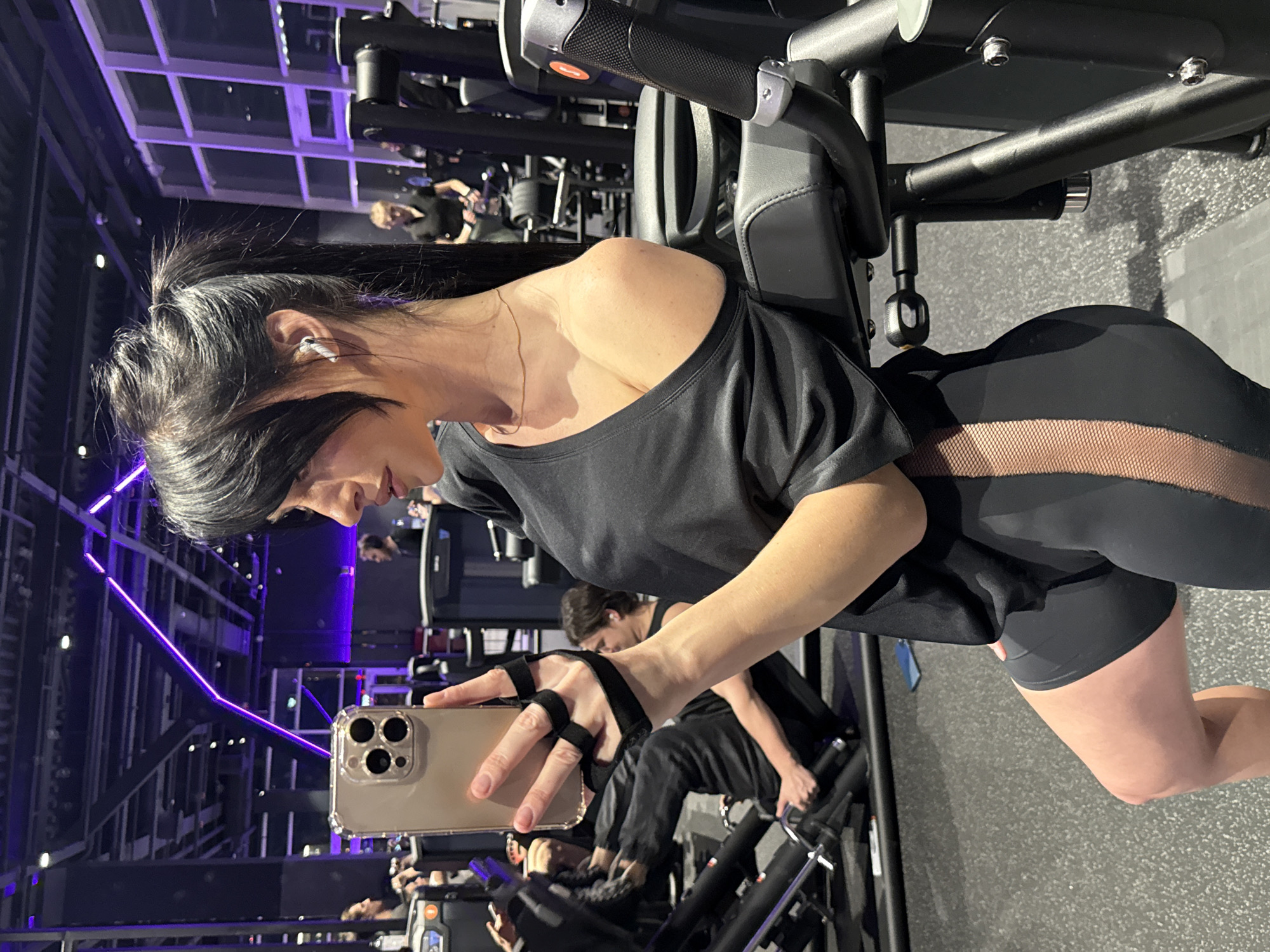 A young woman is captured in a mirror selfie within a modern gym or fitness studio in Krasnoyarsk, Russia. The image is taken from a slightly elevated side angle, with the woman looking down at her gold-colored smartphone, which she holds in her left hand using a black grip strap. She has dark hair styled in a high ponytail and is wearing a black off-the-shoulder top and black athletic leggings with distinctive mesh paneling on the sides, revealing a portion of her skin. White wireless earbuds are visible in her ears, and a delicate gold chain necklace adorns her neck.
The woman appears focused on her phone screen, with a subtle smile on her face, suggesting she is either reviewing or posing for the selfie. She is positioned next to a piece of black gym equipment, possibly an exercise bike or leg press machine, parts of which are visible to her right.
The background reveals a spacious gym environment with other patrons exercising and various fitness machines. Notable are the modern architectural elements of the ceiling, featuring black beams and grid-like structures illuminated by striking purple LED strip lights. These lights cast a vibrant glow, contributing to a contemporary and dynamic atmosphere. The floor is a dark, textured gym surface. No specific time of day is indicated, but the artificial lighting creates an energetic ambiance. There is no discernible text visible in the image.