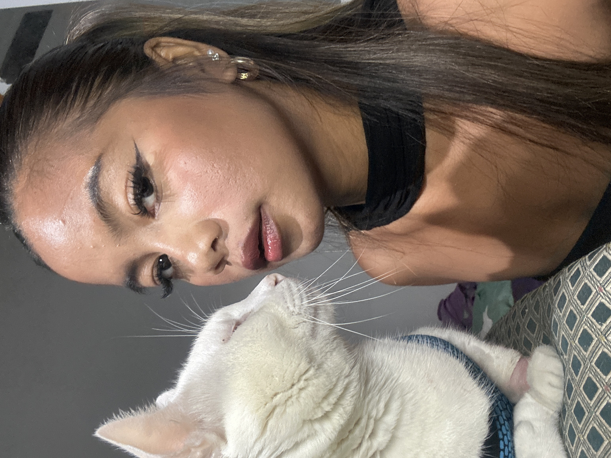 A young woman with long brown hair and makeup is looking at a white cat with blue eyes. The cat is wearing a blue harness. The woman's ear is visible, with a gold hoop earring and a stud earring. She is wearing a black top. The background is blurry. The setting appears to be indoors. There is not enough information in the image to determine the location in North Jakarta, Indonesia.