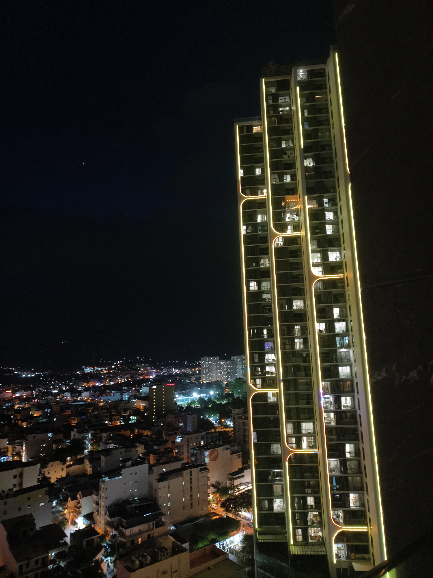 An elevated night view captures a brightly lit skyscraper with yellow and green accent lighting, standing tall amidst a sprawling cityscape. The skyscraper features numerous floors with lit windows, suggesting occupancy. To the left, the urban landscape extends into the distance, characterized by a dense collection of buildings of varying heights, illuminated by a multitude of lights, creating a vibrant tapestry of urban life. The sky above is dark, with only a few faint stars visible, indicating nighttime. The composition is framed by the dark, textured surface of another building or structure on the right side of the image.