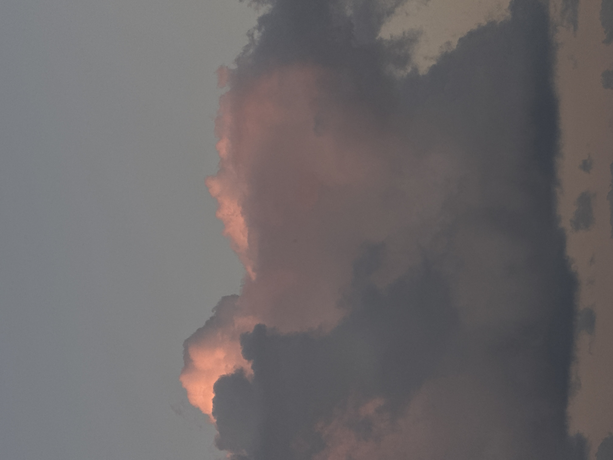 The image is a close-up, vertical shot of cumulonimbus clouds at dusk. The clouds are a mix of dark gray and pink, with the pink hues suggesting the last rays of sunlight. The sky above is a muted, pale gray, indicating a fading light. There are no identifiable people, structures, or geographical features visible, only the sky and clouds. The scene offers no specific information about the city of Denpasar, Indonesia, as it is an entirely natural phenomenon.