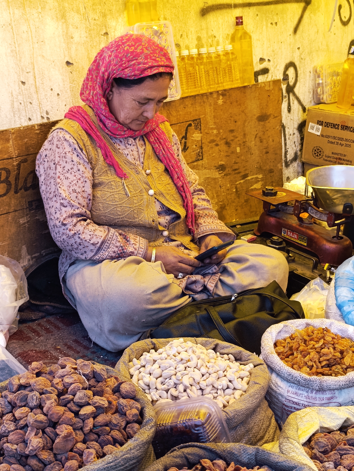 A woman wearing a pink headscarf and a light brown embroidered vest over a patterned long-sleeved shirt sits cross-legged on the ground. She is looking down at a black smartphone in her hands. Around her are several burlap sacks overflowing with dried fruits and nuts, including apricots, raisins, and cashews. To her right, a red and brass weighing scale sits on a surface, and behind her are bottles of yellow liquid and cardboard boxes. The scene appears to be an outdoor market stall, possibly in the city of Leh, India, given the style of dress and the types of goods displayed. The background is a textured wall with some graffiti.