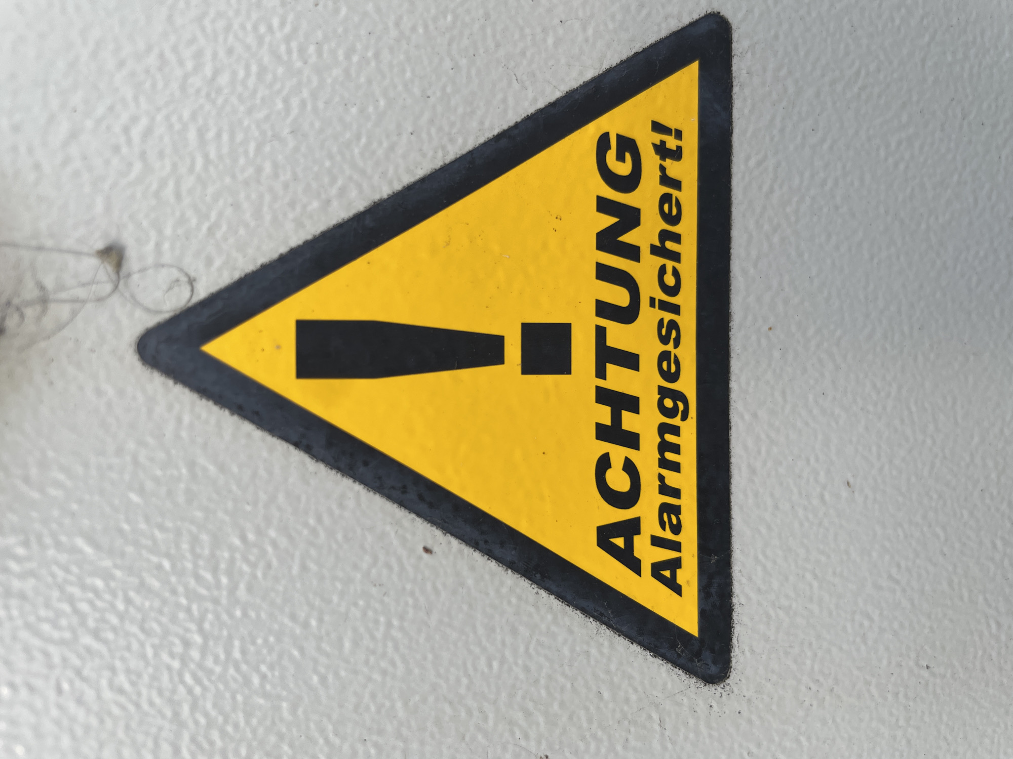 The image shows a close-up of a yellow triangular warning sign with a black border. Inside the triangle, there is a black exclamation mark. Below the exclamation mark, the German word "ACHTUNG" is printed in black capital letters, and below that, the word "Alarmgesichert!" is printed in black. The sign is affixed to a textured, light-colored surface, possibly a metal panel. The background is out of focus and shows no discernible details. There are no people, animals, or any other objects visible in the image. The image does not provide any information about the location in the city of Bad Salzschlirf, Germany.