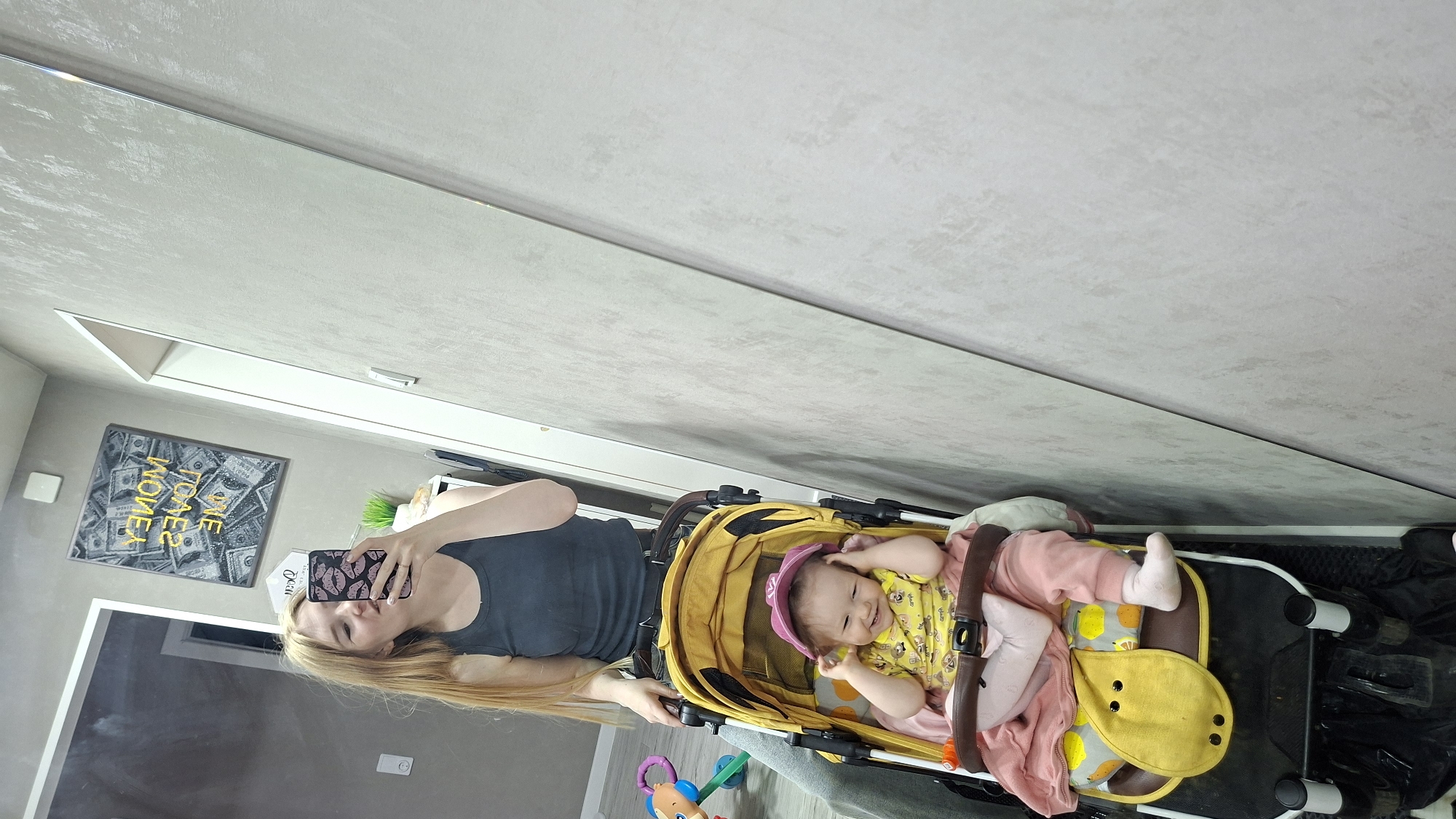 A woman with long blonde hair is taking a selfie in a mirror. She is wearing a dark gray tank top and holding a cell phone with a pink lips pattern on the case. Next to her is a yellow stroller containing a baby wearing a pink hat, yellow shirt, and pink pants. The baby appears to be smiling and interacting with the stroller or her hand. A colorful toy is visible near the stroller. In the background, there's a poster with dollar bills and the word "MONEY" on the wall. The location is implied to be in Chaikovsky, Russia, though specific details to confirm this are absent from the image itself. The setting appears to be indoors, likely a home.