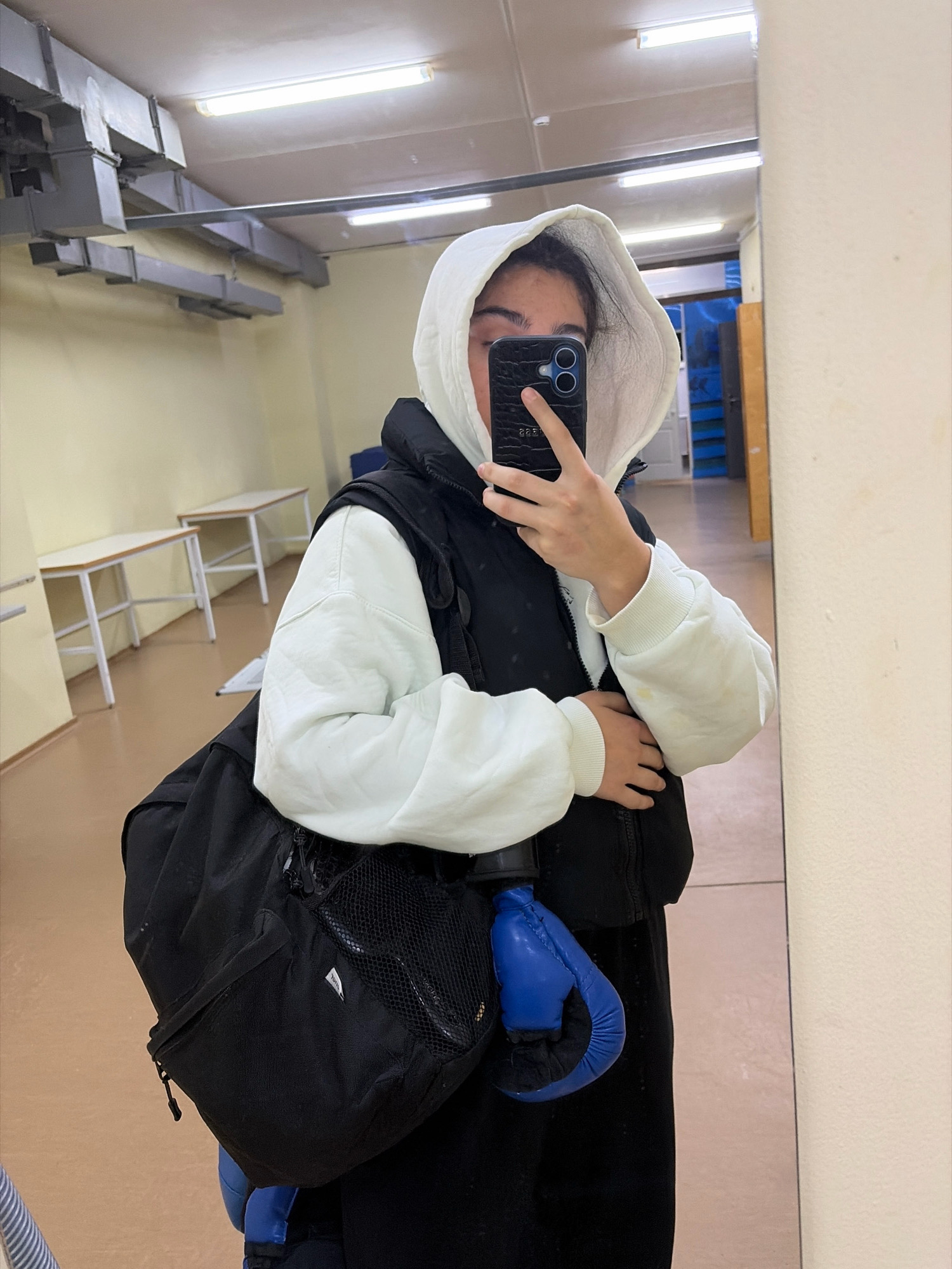A young woman, wearing a white hoodie and a black vest, stands in what appears to be a locker room or gymnasium. She is holding a smartphone in her right hand, taking a mirror selfie. A black duffel bag and a blue boxing glove are visible at her side. The room has a tiled floor and fluorescent lighting. In the background, there are tables and a mirror. The presence of boxing equipment suggests a sports facility, possibly associated with a school or athletic club in Chaikovsky, Russia.