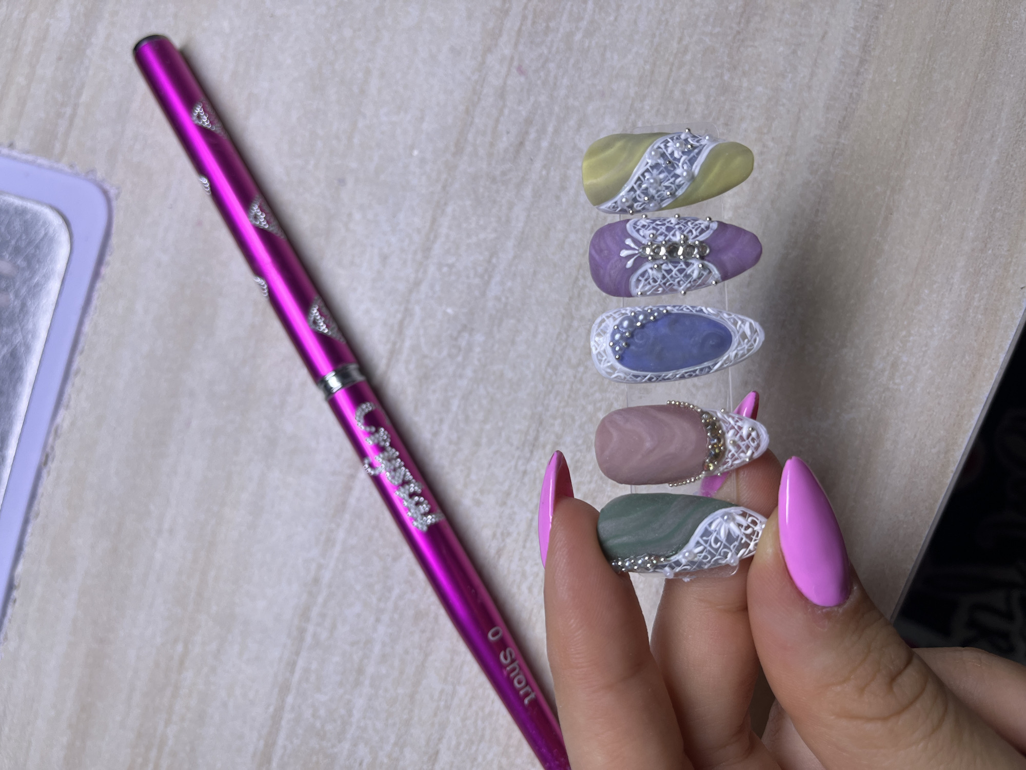 The image shows a hand holding five artificial fingernails with intricate designs. The nails are arranged in a stack, showcasing a variety of colors and patterns. The fingernails display ornate white lace-like patterns with small gemstone or pearl accents. The base colors of the nails vary, including yellow, purple, blue, and green. The hand holding the nails has long, almond-shaped fingernails painted a bright pink color. Next to the hand is a long, pink brush with silver lettering and decorative details. The brush is labeled "0 Short." The background appears to be a light-colored, textured surface. There is nothing in the image to suggest it was taken in Veliko Tarnovo, Bulgaria.