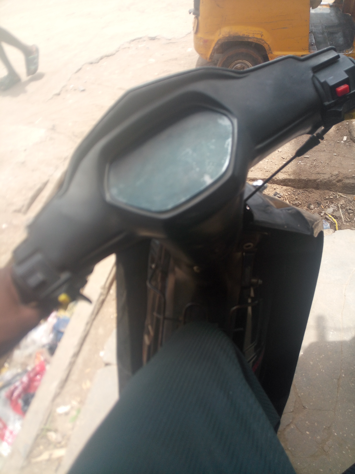 The image shows the dashboard of a black motorcycle. A portion of the operator's leg is visible. In the background is an auto rickshaw. The location appears to be Maiburiji, Nigeria. The surface is dry and likely a road. One other person appears to be walking in the distance, possibly in a sandal.