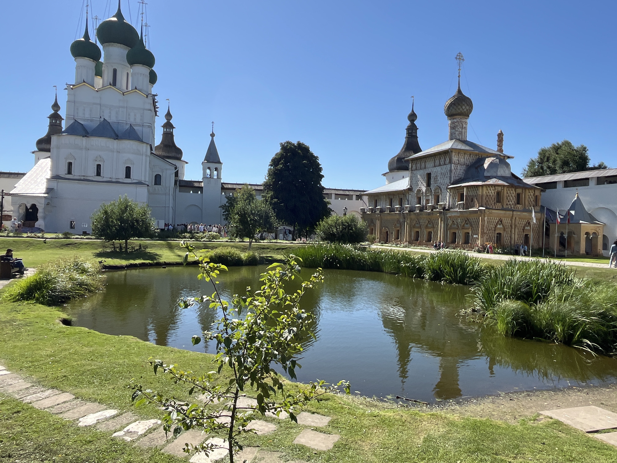 The image shows a bright, sunny day at a historical complex with multiple buildings and a pond. In the foreground, there is a small pond with green water, surrounded by manicured grass. A young tree with green leaves grows on the right side of the pond. Stone pathways are visible on the grass.
In the mid-ground, a large white building with green domes and crosses stands on the left. Its architecture is elaborate, with multiple levels and windows. To the right of the white building, there is a wall and a smaller tower with a grey roof. Further to the right, another ornate building with a patterned facade and a golden dome with a cross is visible. This building has a more intricate design with many windows. The reflection of these buildings can be seen in the pond.
In the background, more of the complex's walls and structures are visible. A line of people can be seen walking in front of the buildings. There are several trees scattered throughout the scene, including a large dark green tree behind the white building and to the left of the ornate building. The sky is clear and blue. The location is identified as Ново-Никольское, Russia.