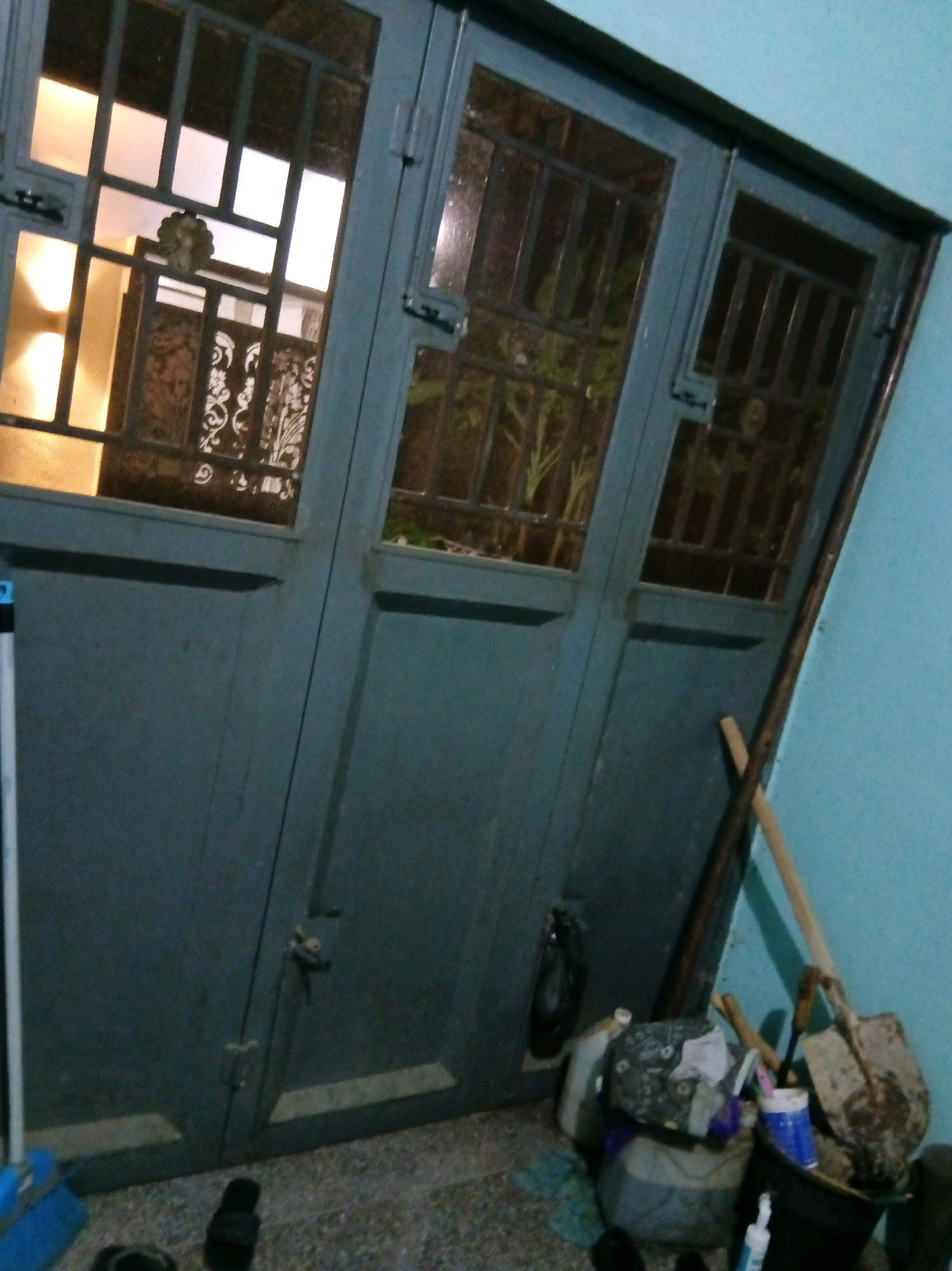 A narrow, angled shot captures a section of a dark gray metal door and the light blue wall it meets in Suka Raya, Indonesia. The door is divided into three vertical panels, each with a grilled window at the top. The left window allows light from within to illuminate ornate metalwork and frosted panels. The middle and right windows are darker, revealing the silhouette of foliage outside. A handle and lock mechanism are visible on the left panel. To the right of the door, leaning against the wall, are gardening tools: a shovel, a rake, and what appears to be a broom handle. Near the base of the door, a black bucket holds various items, including a blue cup and a white bottle. A few dark objects, possibly footwear, are on the floor. A blue mop head with a white handle is positioned to the left of the door.