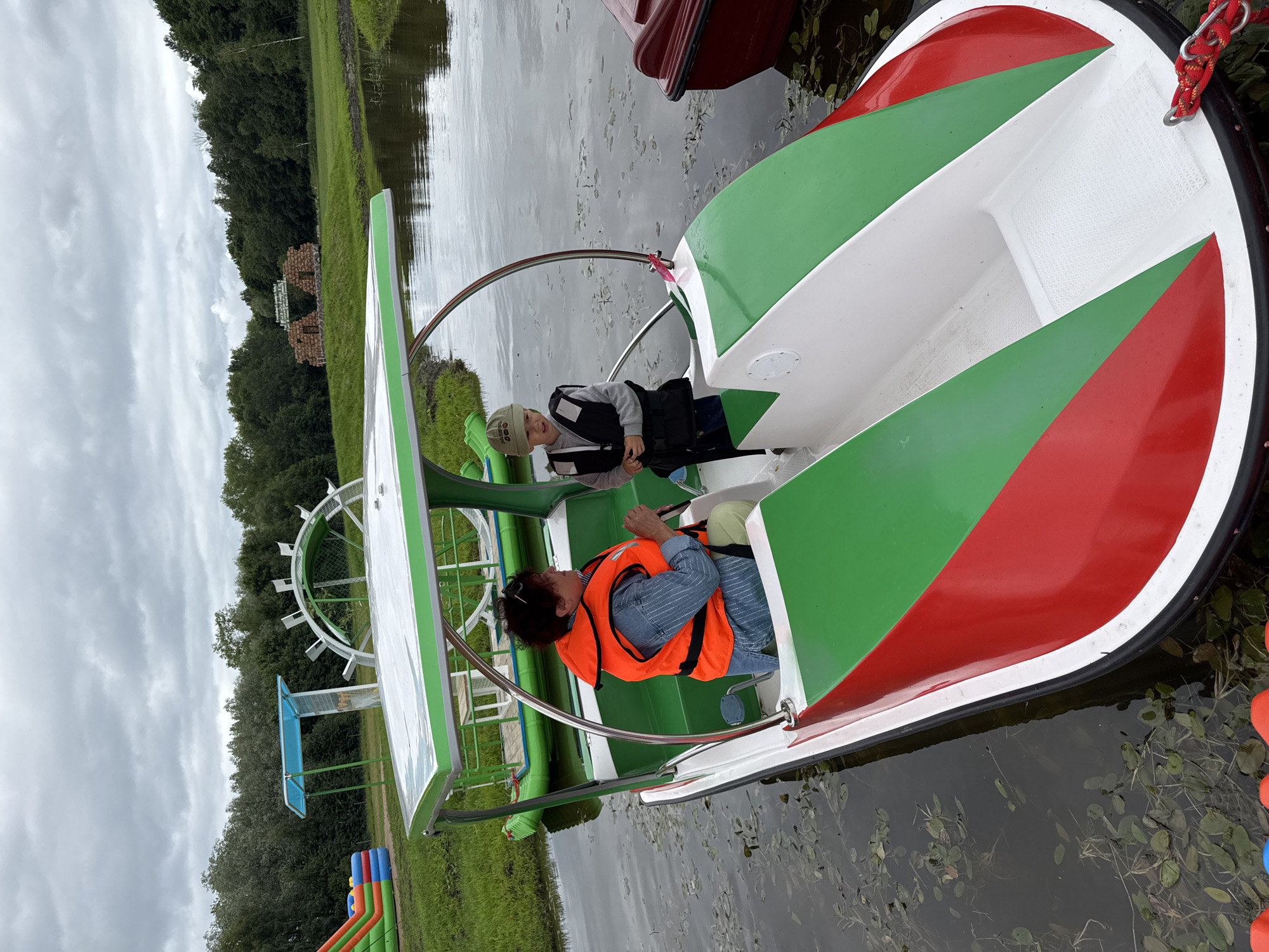 In Bruzdeilynas, Lithuania, a woman wearing an orange life vest and a blue striped shirt is steering a paddle boat. Beside her, a young boy in a grey sweatshirt and a light green cap, also wearing a life vest, stands in the paddle boat. The paddle boat has a white canopy with green trim and red and green accents on its exterior. The boat is on a calm body of water, with lily pads and aquatic plants visible. In the background, a green and inflatable bouncy castle is visible on the grassy shore, along with trees and a cloudy sky. A large, white, circular structure resembling a water wheel is also present in the background. Another red boat is docked to the right of the paddle boat.