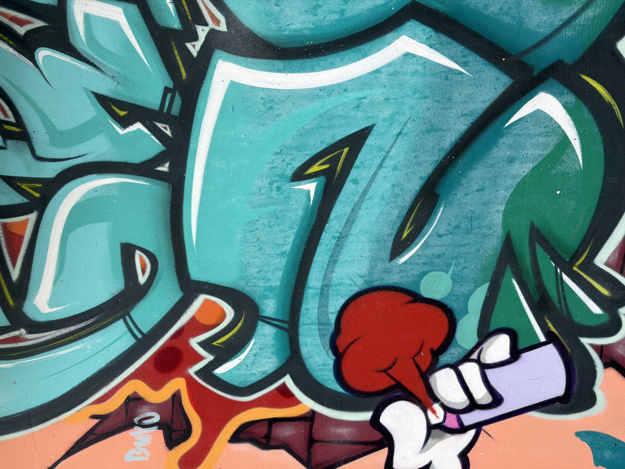A close-up shot depicts graffiti art, featuring an array of vibrant colors, including aqua, red, orange, white, black, yellow, green, peach and lavender. The prominent aqua hues create the background for the abstract lettering and shapes, while the red, orange, and peach tones accent the foreground. A stylized hand holds what appears to be a spray can, releasing a red cloud, indicating a spray-painting action. The surrounding environment appears to be an urban setting, suggested by the graffiti artwork. The location is Varna, Bulgaria.