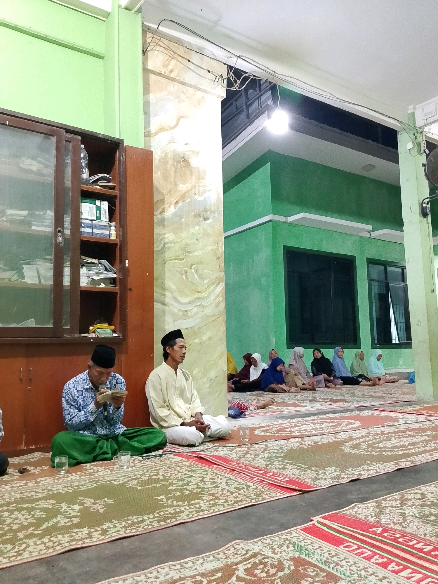 In Kalikondang, Indonesia, two men are seated on the floor in the foreground, facing a bookshelf. The man on the left, wearing a patterned blue shirt and green sarong, is reading from a book. The man on the right, dressed in a light yellow shirt and white pants, is looking forward. Behind them, several women are seated on mats on the floor, some wearing hijabs. The environment appears to be an indoor space with green walls and a white ceiling, illuminated by artificial light. A large wooden bookshelf with glass doors is visible on the left, containing books and papers. The floor is covered with patterned mats. The scene suggests a religious gathering or study session.