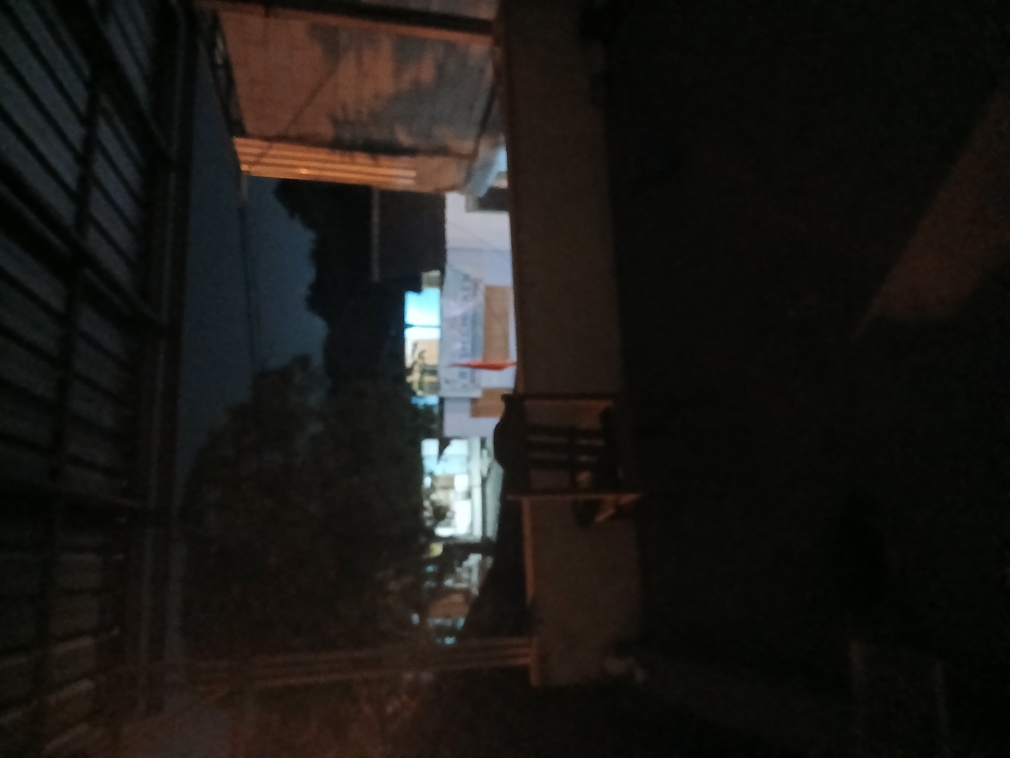 The image depicts a dimly lit outdoor scene, possibly a patio or rooftop, under a corrugated metal roof. In the background, there are buildings with some lights on, suggesting it is nighttime. A single wooden chair is positioned in the middle ground. An orange flag is visible on a pole to the left of the chair. The overall impression is one of an abandoned or quiet outdoor space at night. There is no indication of the specific location within Tenggarang, Indonesia.