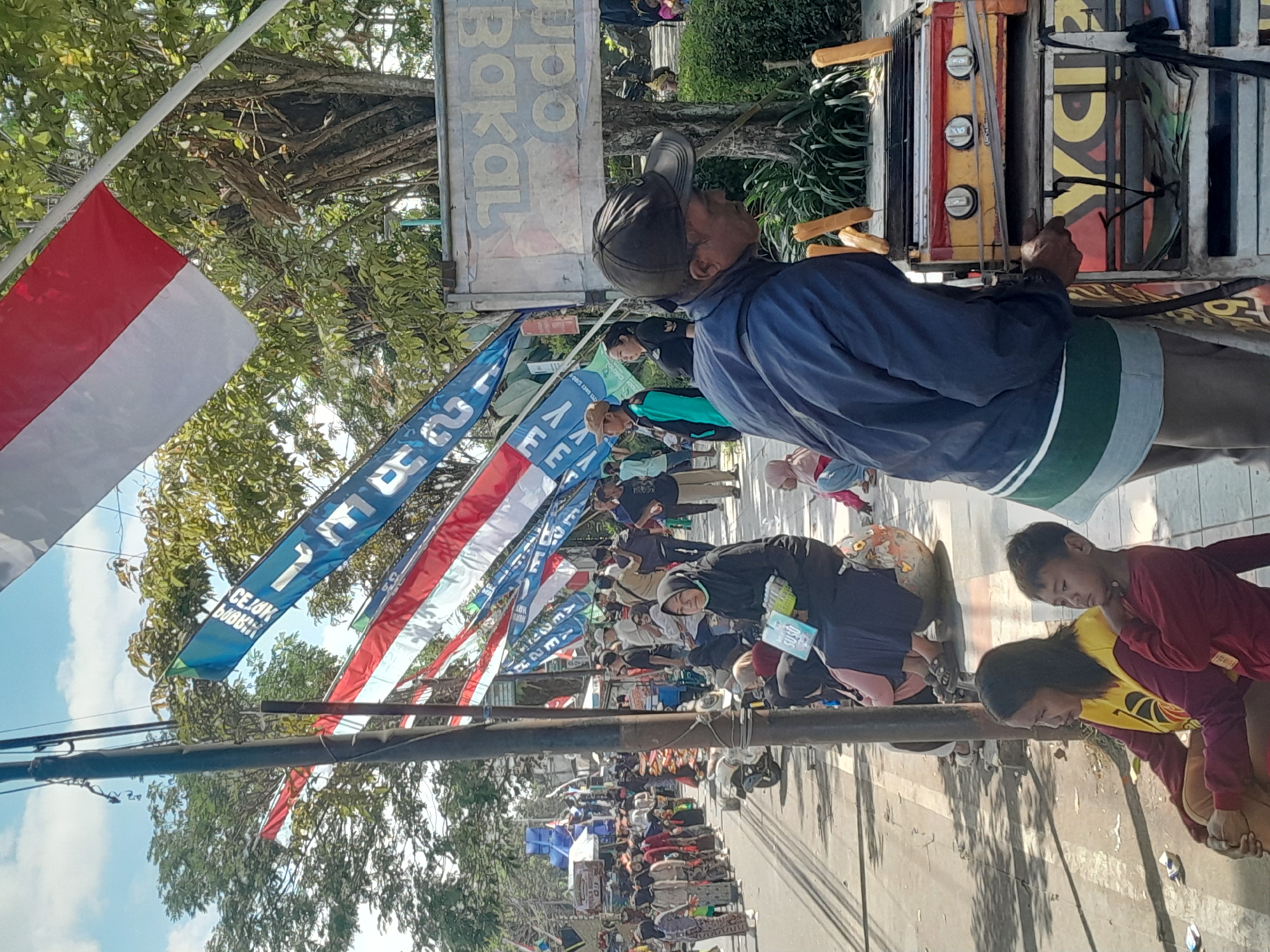 In Genteng Kulon, Indonesia, on a sunny day, a man wearing a blue jacket and a dark baseball cap stands behind a food cart. The cart has a yellow and red grill with three burners and a metal grate. Hot dogs or sausages are seen on the grill. Beneath the grill, a display with "YUCI" written in large yellow letters is visible, along with what appear to be stacks of paper or promotional materials. The man's left hand is on the cart.
To the left of the food cart, two children sit on the ground. A woman with dark hair, wearing a yellow shirt with a red neckline and a maroon long-sleeved shirt, is seated with her right arm around the shoulder of a young boy in a red long-sleeved shirt. The boy is looking towards the right with his chin resting on his arm. A second child, a young girl wearing a pink headscarf and blue pants, is also visible.
In the background, a busy street scene unfolds. A crowd of people, many wearing traditional Indonesian attire, are lining the sidewalk and street. Some are walking, while others are gathered in groups. Several flags, including red and white Indonesian flags, are strung across the street, along with blue banners with white lettering. A motorcycle is parked on the side of the road. The environment appears to be an outdoor market or a street fair, with trees and buildings forming the backdrop.