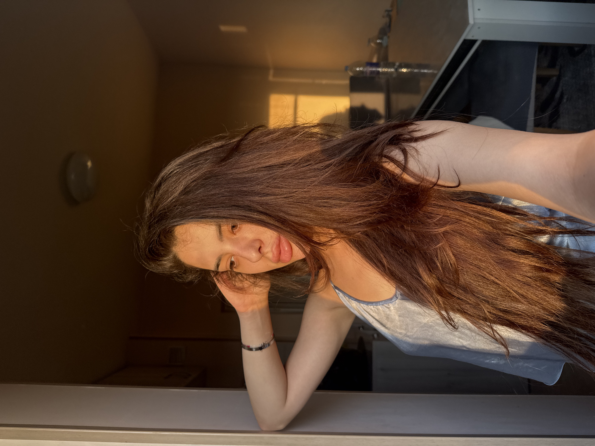 The image displays a young woman in an indoor setting, illuminated by strong, warm golden light entering from the right, likely natural sunlight during golden hour. She is positioned in the left-center, looking towards the right with a relaxed expression. She has long, dark brown hair that cascades over her left shoulder and chest. Her left arm is bent, with her hand touching the back of her head, and her elbow resting against a light-colored door frame or wall on the left side of the frame. She wears a light blue or silver-grey spaghetti strap top, and a silver-toned bracelet is visible on her left wrist. The background on the right is darker, revealing a ceiling light fixture in the upper portion and a reflective surface, possibly a table, extending from the mid-ground. A clear plastic water bottle and a glass are situated on this surface. A rectangular patch of golden light is cast on a far wall, alongside several shadows, one of which resembles a human silhouette. The specific location in Plovdiv, Bulgaria, cannot be ascertained from the visual information provided.