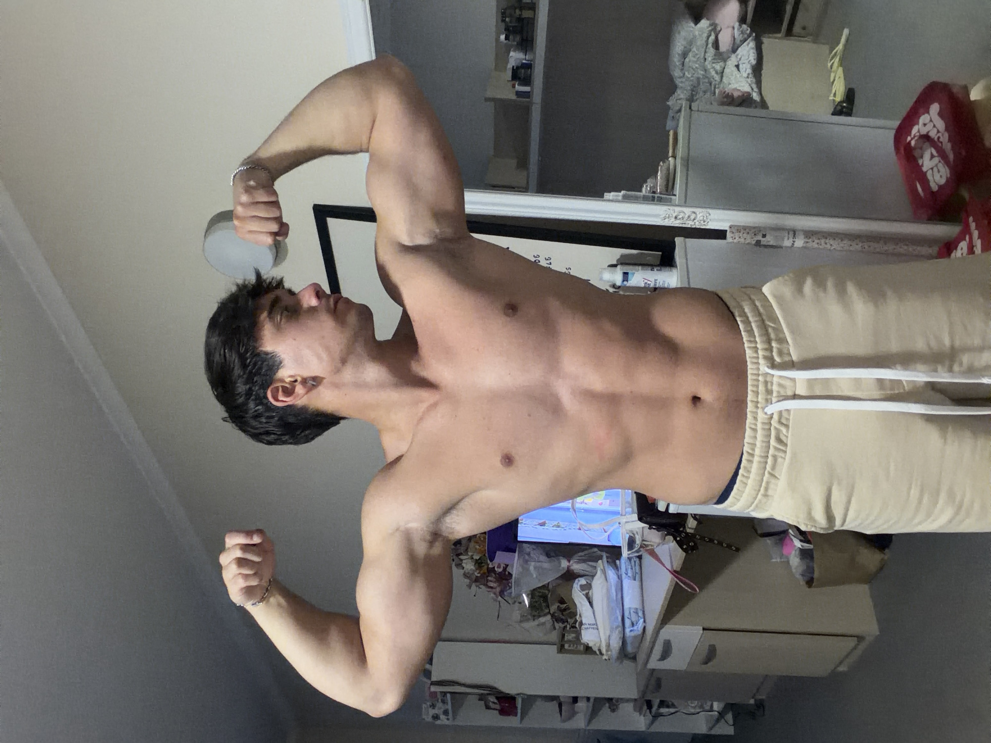 The image, originally rotated 90 degrees counter-clockwise, captures a young man flexing his muscles in what appears to be a personal room, likely in Sofia, Bulgaria. He is shirtless, displaying a lean and muscular physique with defined biceps, chest, and abdominal muscles. He wears light-colored (beige or khaki) sweatpants with white drawstrings and a silver bracelet on his left wrist. His left hand holds a small, circular grey object near his head, while his right fist is clenched. He poses, looking upwards and to his left, appearing confident.
The background suggests an indoor setting with some clutter. To the man's right, a white dresser or cabinet holds various items, including bottles, fabric, and a red object with the word "CASA" partially visible. A mirror reflects shelves filled with bottles, and a whiteboard or framed picture with indistinct writing is visible behind his right shoulder. Further right and lower, another surface is cluttered with items, and a screen (possibly a TV or monitor) displaying colorful content can be partially seen. The room is well-lit, suggesting daytime or bright interior illumination.