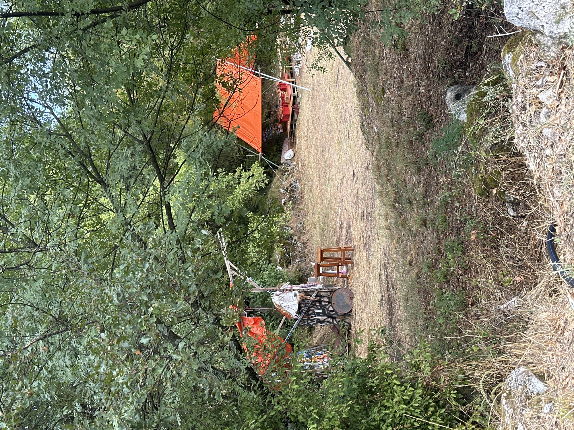 The image depicts an outdoor scene likely within a wooded area, possibly on the outskirts of Cetinje, Montenegro. In the foreground, dry grasses and sparse vegetation cover the ground, interspersed with rocks. Dominating the middle ground is a clearing with a dirt or gravel surface.

To the left of the clearing, a temporary structure is visible, draped with an orange tarp and other materials, possibly for shelter or shade. Various objects, including what appear to be drums and wooden stools, are arranged around this structure.

Further into the clearing and to the right, a larger orange tarp is erected on poles, creating a makeshift canopy. Beneath this tarp, a raised wooden platform is visible, supporting several red crates or boxes. The surrounding area shows signs of disarray, with scattered debris and other undefined objects. The entire scene is framed by dense green foliage from trees and bushes, suggesting a natural or semi-wild environment. No individuals are present in the image.