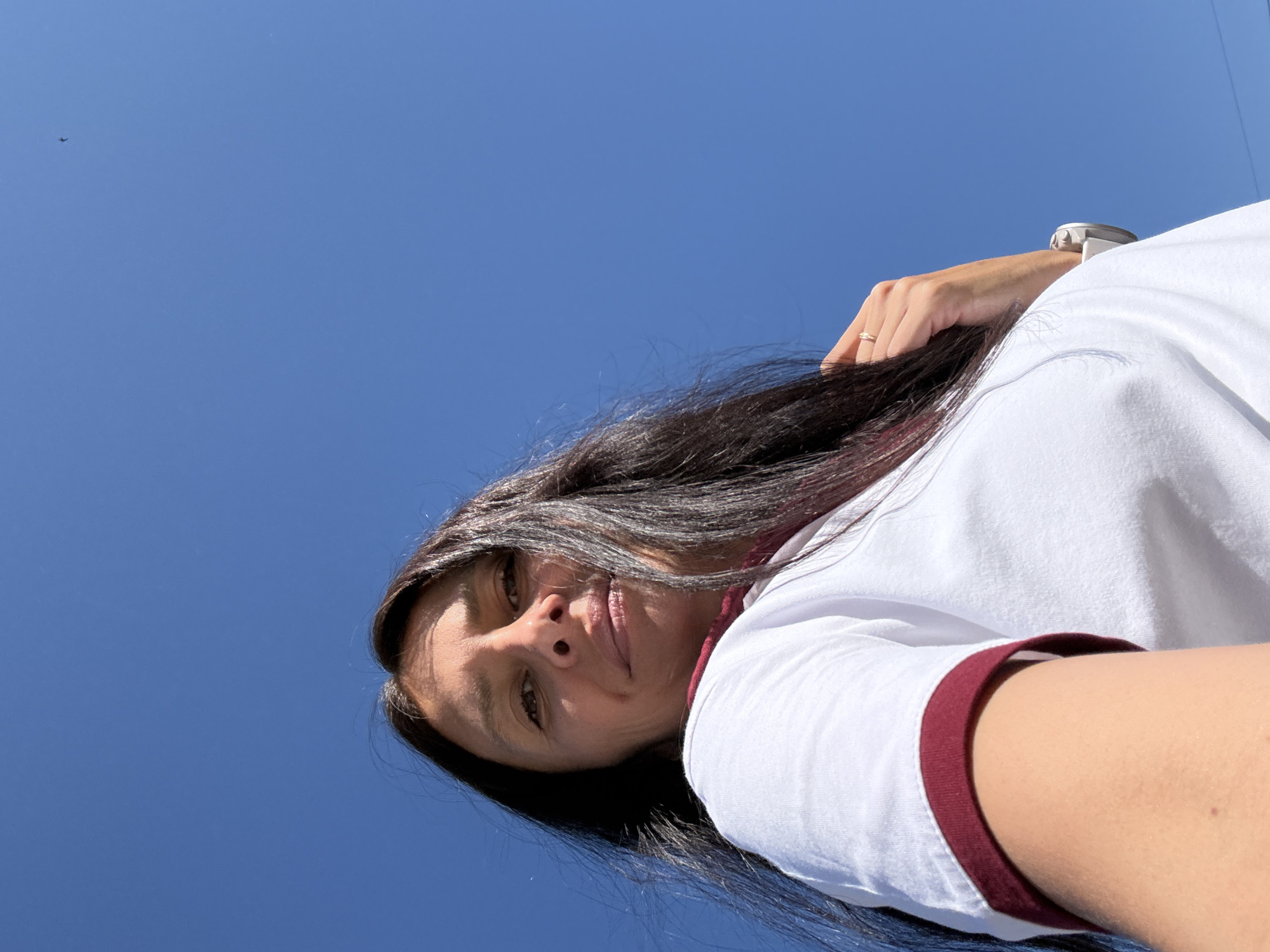 The image is a low-angle, close-up selfie of a woman against a clear blue sky. The woman has long, dark hair, which is partially obscuring her face. She is wearing a white t-shirt with maroon trim around the collar and sleeves. Her left arm is bent, and her hand, adorned with a ring, rests near her head. A small, dark object, possibly a bird or an insect, is visible in the upper right portion of the sky. The overall scene suggests an outdoor setting on a sunny day. The location is identified as Chaikovsky, Russia.