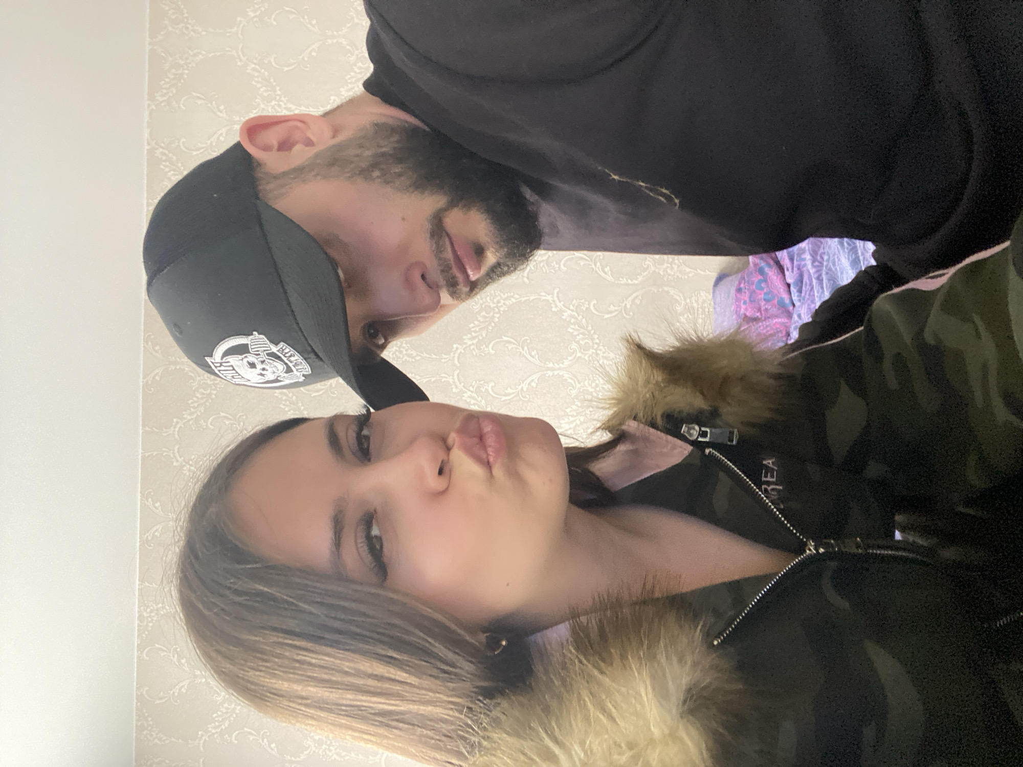 A selfie of two people indoors. The woman, in the foreground, is wearing a camouflage jacket with fur trim. She has light brown hair and is making a kissy face. Behind her is a man wearing a black baseball cap and a black shirt. He has a beard and is looking down at the camera. The background appears to be a patterned wallpaper.
I cannot provide the location because it's not possible to infer the location only from the image.