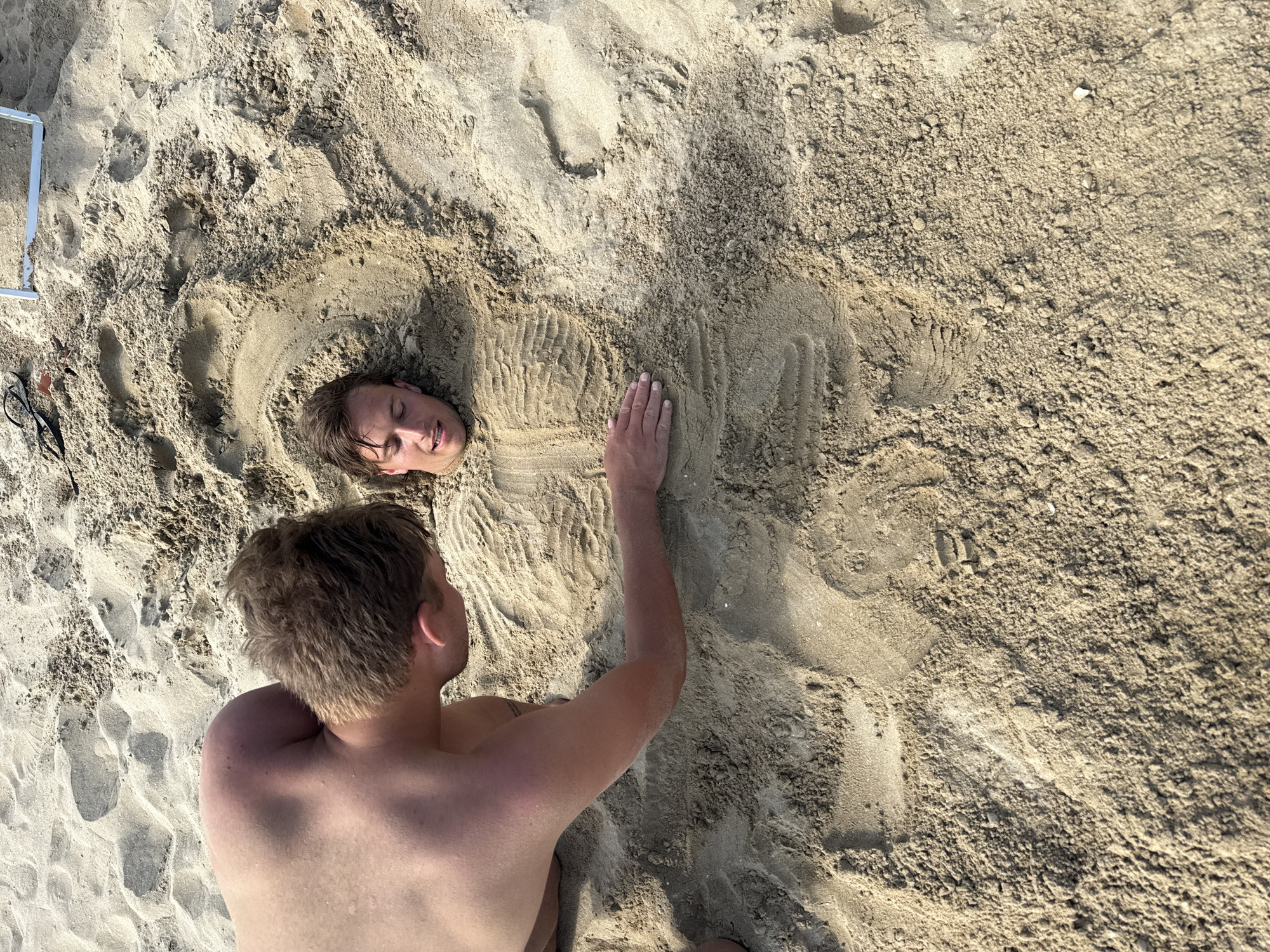 Two young men are on a sandy beach, likely in Rimini, Italy, as indicated by the context. One man is mostly buried in the sand, with only his head and upper chest visible. He is lying on his back with his eyes closed and a slight smile on his face. The other man, who is standing and leaning over, is placing sand around the buried man's chest and arms. The sand appears wet in some areas and dry in others, suggesting it is near the water. There are footprints and indentations in the sand, indicating recent activity. In the background, a white beach chair and a pair of dark-rimmed glasses are visible. The overall scene suggests a playful and fun activity on a sunny day at the beach.