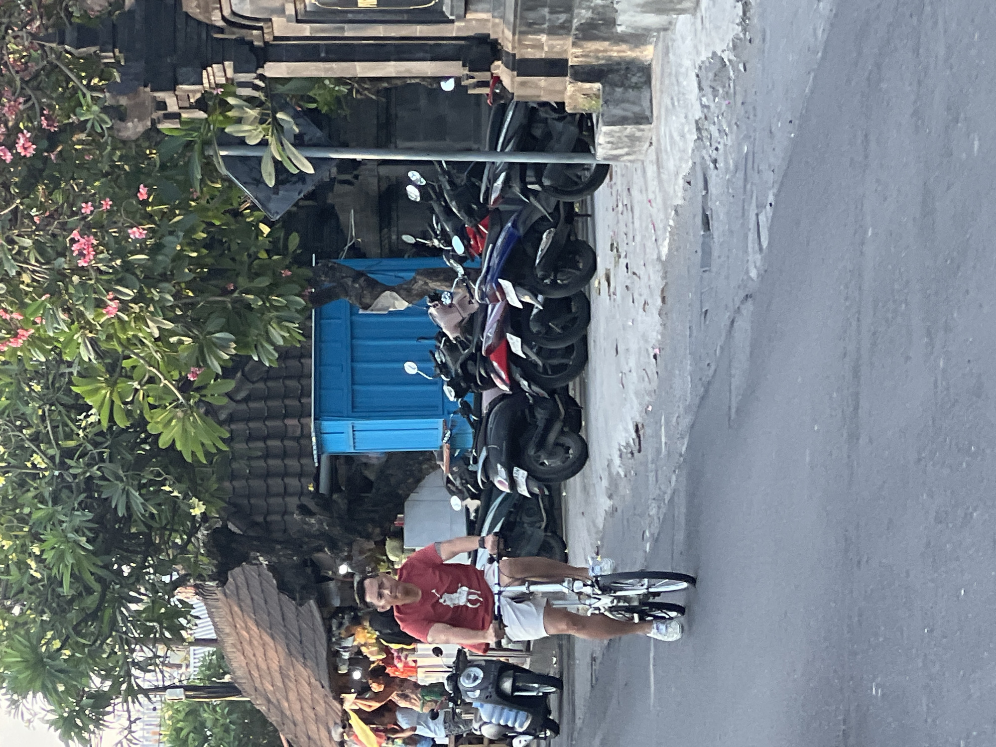 A man rides a foldable bicycle down a paved road in Denpasar, Indonesia, wearing a red t-shirt with a polo player graphic and white shorts. He is in motion, with his legs pedaling. The road is lined with parked motor scooters on the right side. Behind the scooters, there is a building with a blue facade and a tiled roof. Trees with green foliage and pink flowers overhang the scene. The street appears to be a commercial area, with stalls or shops visible in the background. The time of day appears to be late afternoon or early evening, given the lighting. The overall atmosphere is casual and urban.