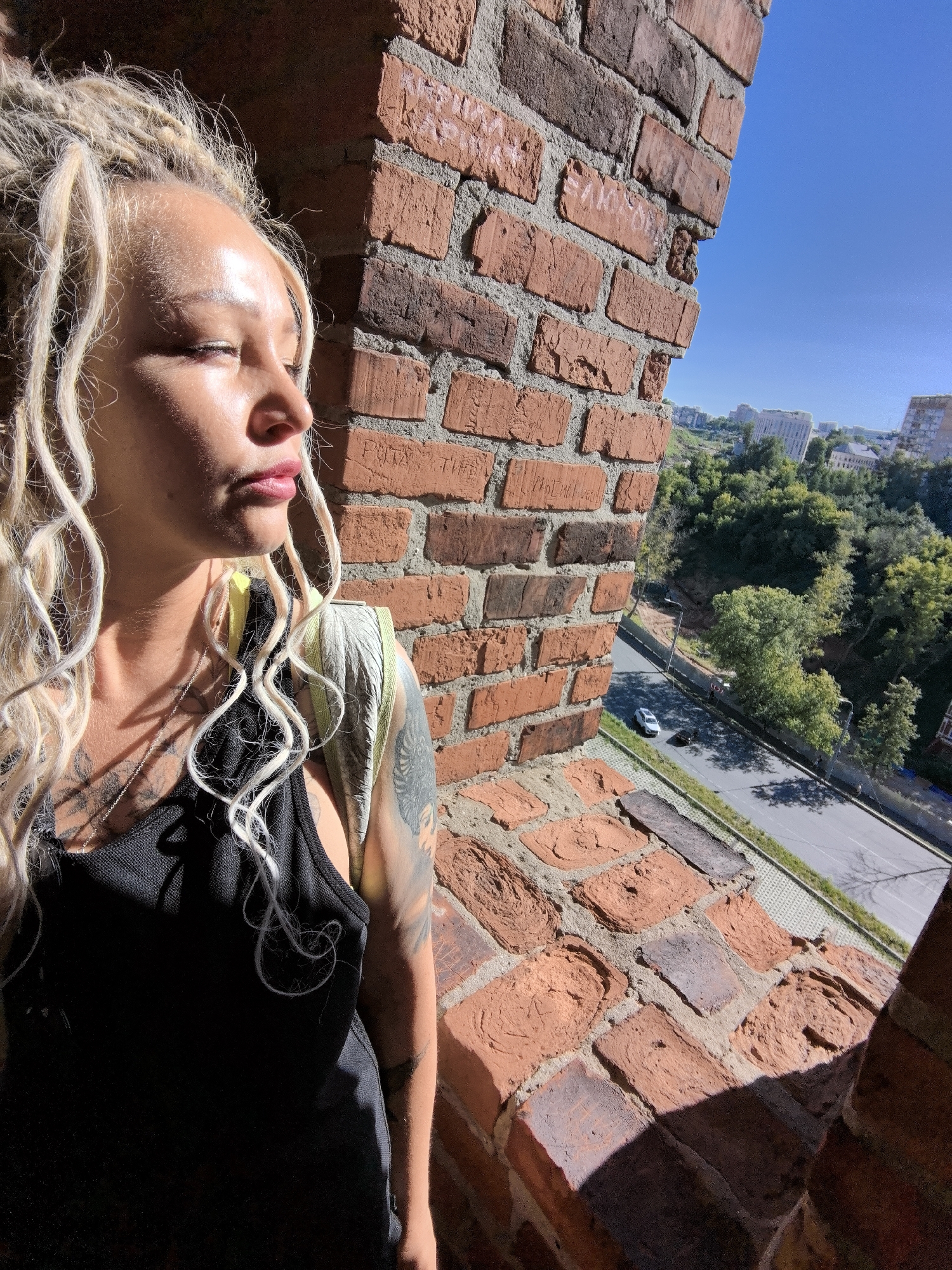 This is a medium-shot, eye-level, outdoor, daylight photograph featuring a woman with blonde dreadlocks and tattoos standing next to a brick wall. The woman is in the foreground on the left, with her face turned to the right and her eyes closed. She is wearing a black tank top and a silver necklace. The brick wall is to her right, extending into the midground. Sunlight is casting shadows across her face and the wall.
In the background, a tree-lined street with two cars is visible, and beyond that, there are buildings and a bright blue sky. The overall scene suggests an urban environment. Based on the architecture and landscape, the location is likely in the area of the Nizhny Novgorod Kremlin in Nizhny Novgorod, Russia, specifically overlooking the Kremlin embankment and the Volga River.