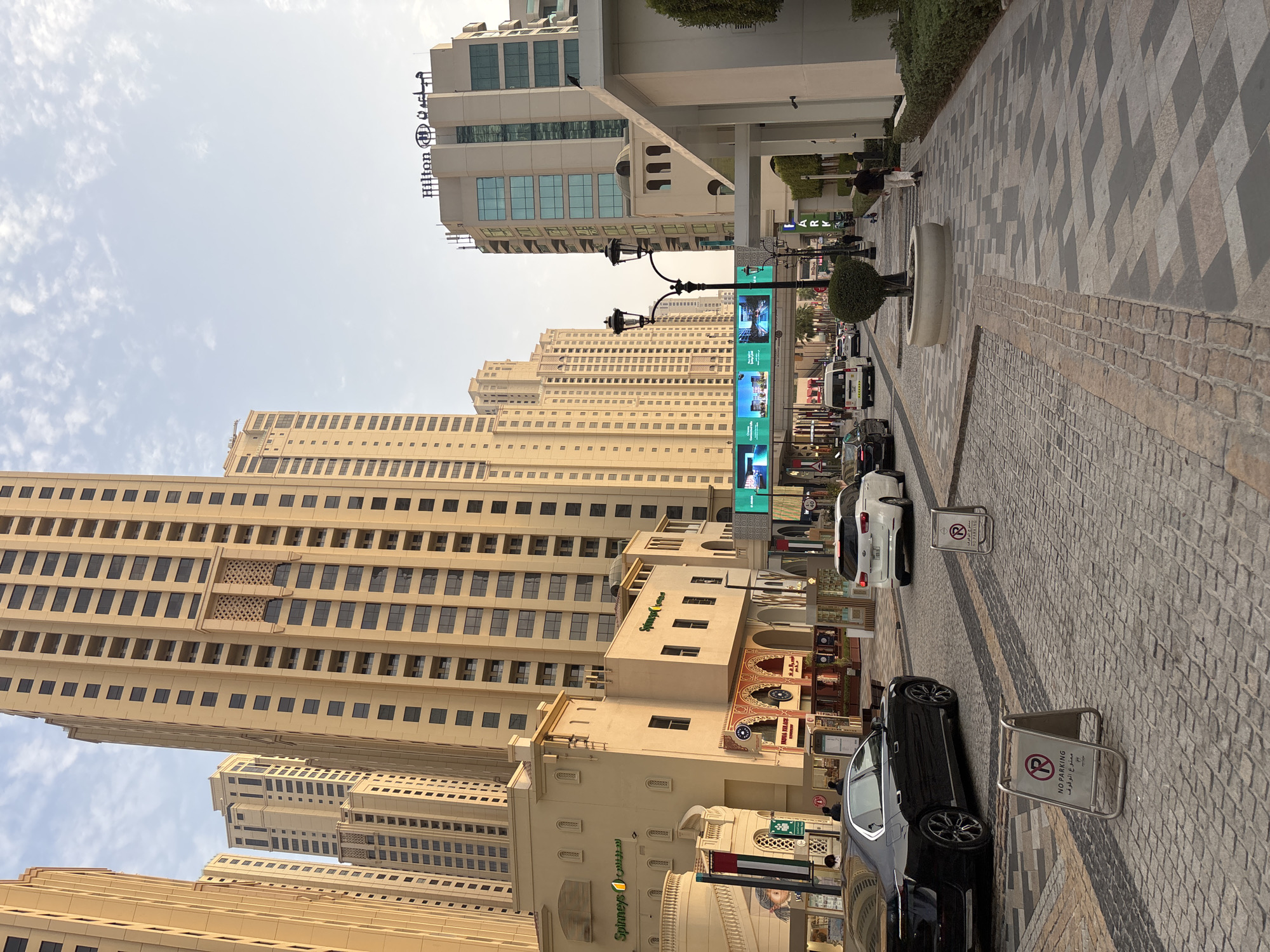 The image depicts a street scene in Dubai, United Arab Emirates, likely in the Jumeirah Beach Residence (JBR) area given the architecture and the presence of the Hilton Jumeirah Beach Resort sign. Tall, modern residential buildings with beige facades and numerous windows dominate the background. In the foreground, a paved street is lined with shops and restaurants. Several cars, including a black sedan and a white SUV, are visible on the street. Two "No Parking" signs with Arabic text are placed on the sidewalk. Pedestrians are sparsely scattered along the walkway. A large digital billboard displays advertisements for what appears to be a resort or residential development. The sky is clear with some wispy clouds. The overall impression is one of an urban, upscale commercial and residential district.