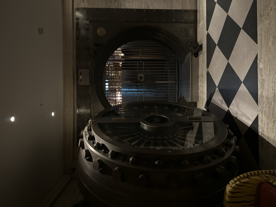An interior photograph captures the open, imposing circular vault door of a former bank, located at The Ned on Poultry, London, United Kingdom. The massive, dark grey metallic vault door, featuring prominent bolts around its circumference, is ajar on the left, revealing internal mechanical components. This vault is set within a thick, riveted metal frame, integrated into a light-colored, textured stone wall. Above the circular vault opening, a small rectangular control panel with three distinct colored indicators (red, white, and grey/blue) is affixed.
Beyond the primary vault door, a secondary, vertically barred metal security gate stands open, providing a view into a dimly lit interior space characterized by further metal grates or shelving. The foreground floor is covered with a black and white checkerboard tile pattern, which transitions to a lighter, possibly stone, floor closer to the vault entrance. The white ceiling above features multiple recessed spotlights. A portion of a yellow and black striped armchair or sofa is visible in the lower-left corner of the image. No individuals are present, and no discernible actions or interactions are occurring.
