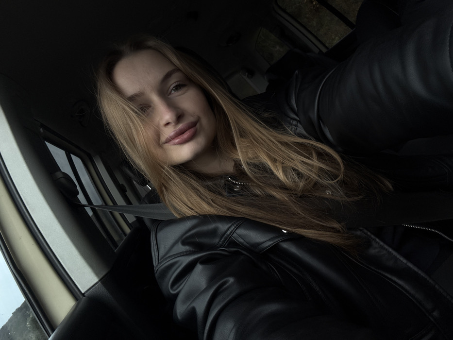 The scene is set within the passenger compartment of a vehicle, located in Brežine, Montenegro. A single female individual, appearing young adult, is centered in the frame, taking a self-portrait. She has long, light brown hair, parted on her left, with strands partially obscuring the left side of her face. Her lips are full, and her mouth exhibits a slight upward curve at the corners. The subject wears a black leather jacket over a dark, possibly zipped, inner garment. A dark gray seatbelt is visible, crossing diagonally from the upper left of the frame over her left shoulder towards her chest area. The vehicle's interior features a light-colored ceiling liner, a visible sunroof mechanism, and dark trim elements. Through the vehicle's windows, obscured outdoor elements are visible, including distant, possibly elevated, landforms and blurred vegetation, suggesting an overcast sky.