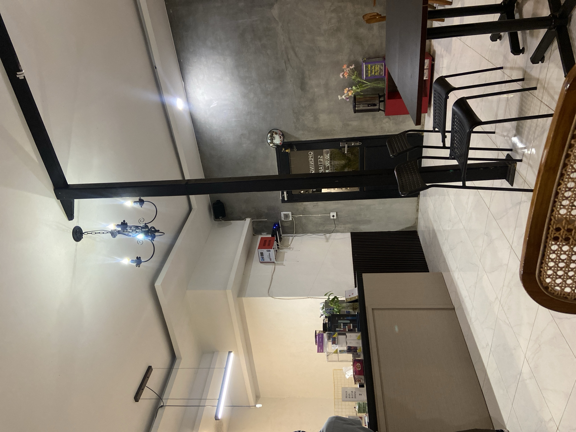 The image depicts the interior of a modern cafe, likely in Sekar Mawar, Indonesia. The space features a high ceiling with a modern pendant light and a traditional chandelier. A long, grey counter runs along the left side of the room, behind which is a wall with shelves holding various products, including boxes and a plant in a vase. To the right, a dark grey, textured wall serves as a backdrop for a black door with glass panels and a circular mirror above it. In front of this wall, there is a bright red cabinet topped with a stainless steel dispenser and a vase of flowers. A black table with chairs is positioned in the foreground to the right, set against a polished tiled floor. The overall ambiance is contemporary with elements of industrial design. There are no visible people actively engaged in actions, though the setup suggests a functional cafe environment.