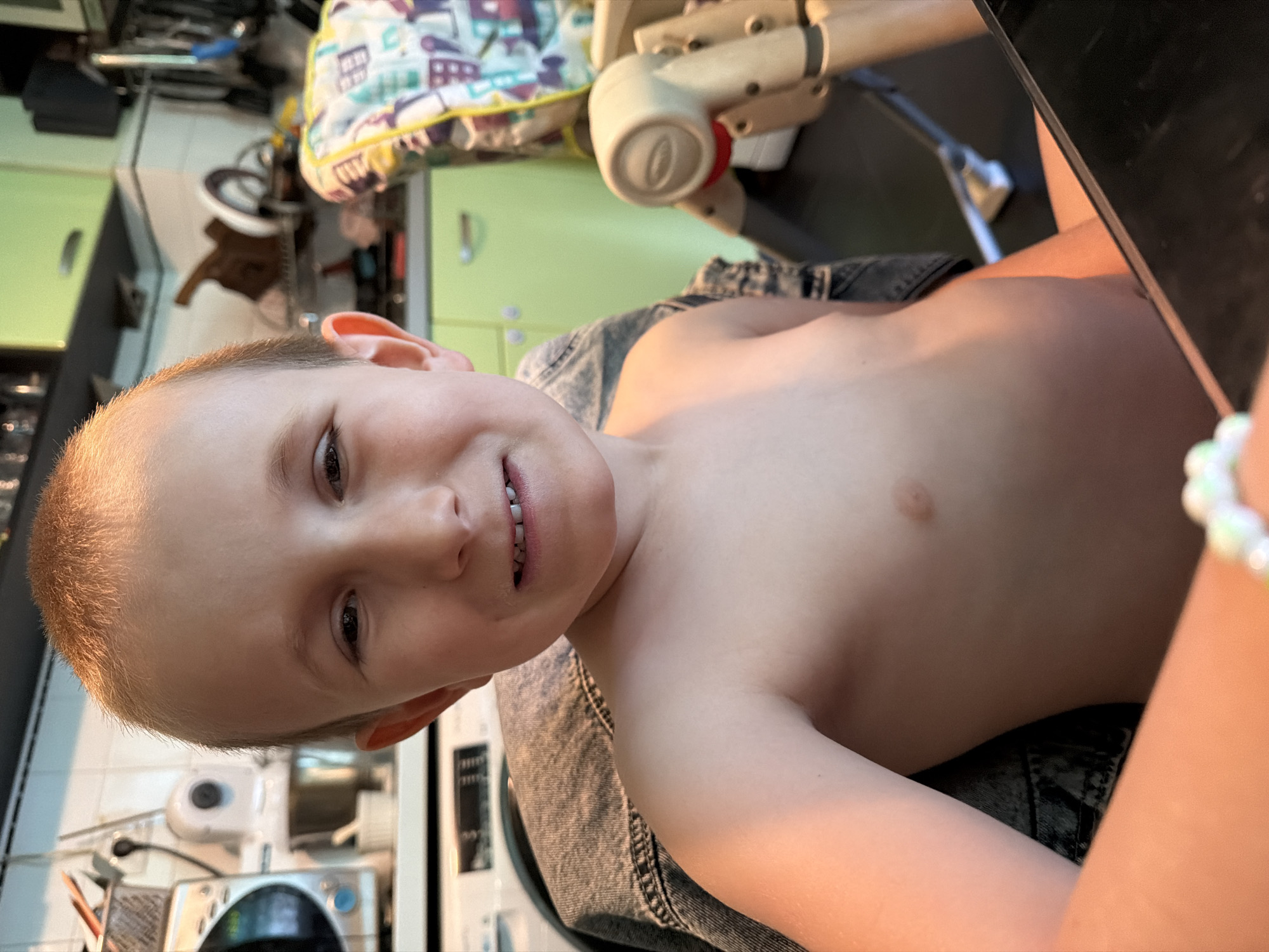 The image is a close-up, low-angle shot of a young boy, likely between 4 and 6 years old, with short, light brown hair and fair skin. He is shirtless, wearing denim shorts. The boy is looking directly at the camera with a playful, slightly toothy smile. His right eye is visible, and his mouth is open, revealing his teeth. The lighting suggests it is daytime, with warm sunlight illuminating his skin.
In the background, the scene appears to be a domestic setting, likely a kitchen or utility area, given the presence of what looks like kitchen cabinets and appliances. There is a colorful, patterned fabric object, possibly a baby carrier or blanket, in the upper background. A light-colored mechanical arm or structure, perhaps part of a baby bouncer or walker, is also visible. The overall impression is casual and intimate, capturing a candid moment. The boy's expression conveys a sense of happiness and engagement. There is no visible text in the image. The location context is Sofia, Bulgaria, but this cannot be discerned from the image itself.