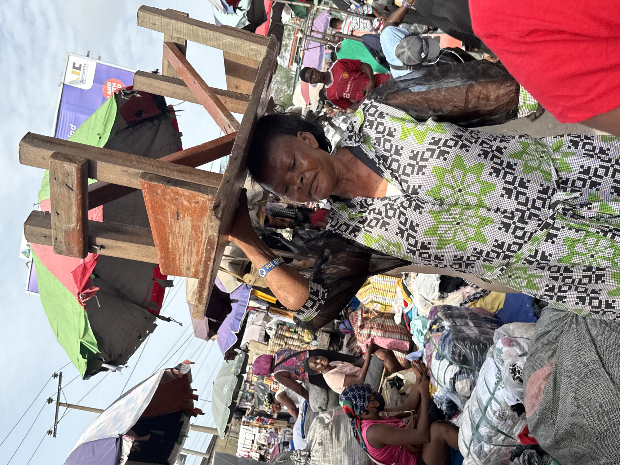 This media file captures a vibrant and crowded outdoor market scene in Lekki, Nigeria, during what appears to be daytime under a partly cloudy sky.
The central subject is an older Black woman seen in a profile view, carrying a large, heavy-looking wooden structure—possibly a disassembled market stall or display frame—on her head. Her eyes are closed, and her facial expression suggests concentration, effort, or perhaps a moment of brief rest amidst the bustling environment. She is dressed in a distinctive patterned gown featuring green, white, and black geometric designs.
Around the woman, the market is teeming with activity. Numerous piles and bales of clothing and textiles are stacked high, indicating a strong focus on fabric or second-hand goods. Various colorful umbrellas provide shade over different market stalls. In the background, other individuals are visible; some are standing, while others are seated among their wares. One person can be seen wearing a red jersey with the "TeamViewer" sponsor logo and "Manchester United" branding. A blue wristband on an arm in the mid-ground shows the partially visible text "with You."
Above the market, a large billboard is partially visible against the sky, displaying commercial text such as "Jerclems," "AND DEO," and "ind." Utility poles and wires are also discernible in the upper left portion of the image. The overall impression is one of a busy, dynamic commercial hub, characteristic of a typical market in the region.