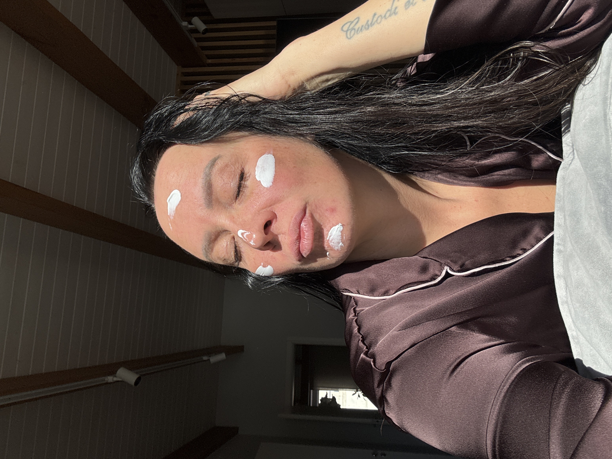 A woman with dark hair and fair skin is lying down, facing the camera. She has several dabs of white cream or lotion on her forehead, cheeks, and chin. She is wearing a brown satin pajama top with white piping. Her left arm is raised, with a tattoo visible on the inner forearm. The background includes a ceiling with exposed wooden beams and a dark wall with a window in the distance. The location is not discernible from the image alone.