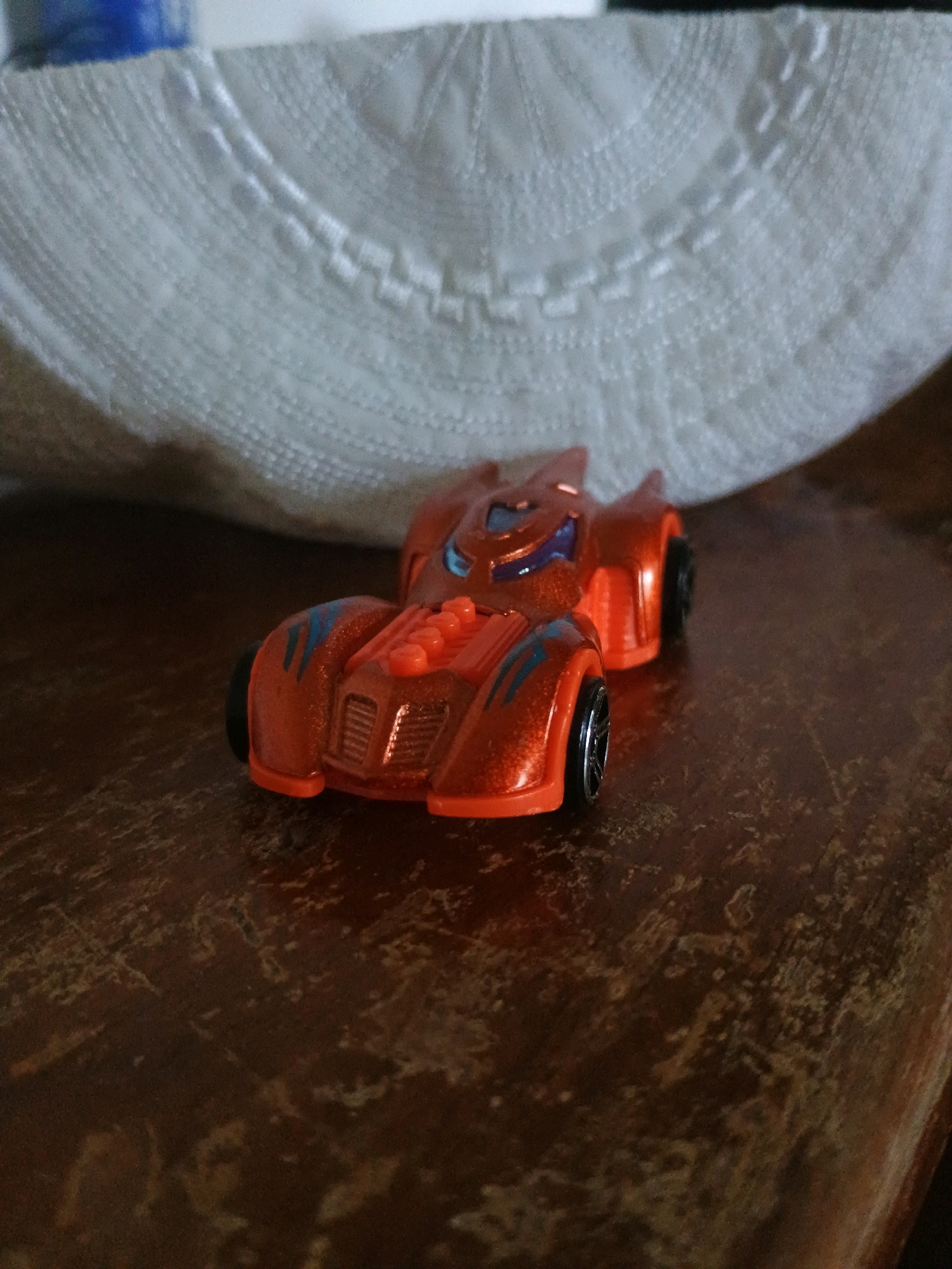 An orange toy car, designed in a futuristic style with blue accents, rests on a dark wooden surface. Behind the toy car, a white, textured fabric is visible, with a blurred blue object in the upper left corner. The scene appears to be indoors, and there is no indication of the city of Tirur, India, or any specific location within it.