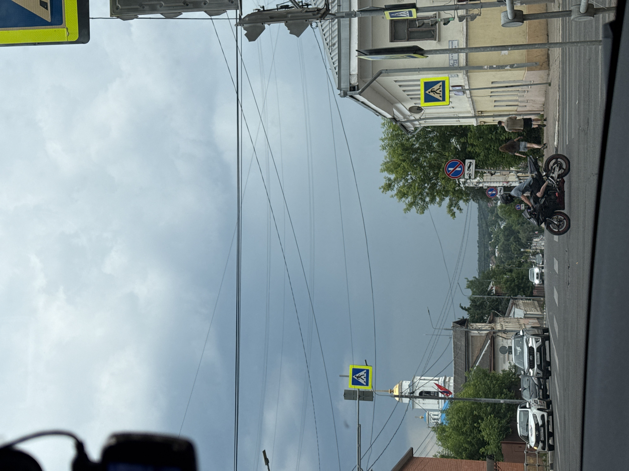 The image shows a street scene in Serpukhov, Russia, on a cloudy day. In the foreground, a black motorcycle with a rider wearing a helmet is in motion, crossing a pedestrian intersection. To the right, two pedestrians are walking away from the camera on a sidewalk. Behind them, a light-colored building with a yellow facade stands on a corner. Further down the street, a white SUV and a white sedan are stopped at a crosswalk. Across the street, a church with a blue dome and a white steeple is visible. Overhead, multiple power lines crisscross the sky, and traffic signals and pedestrian crossing signs are present. The sky is filled with dark, heavy clouds.