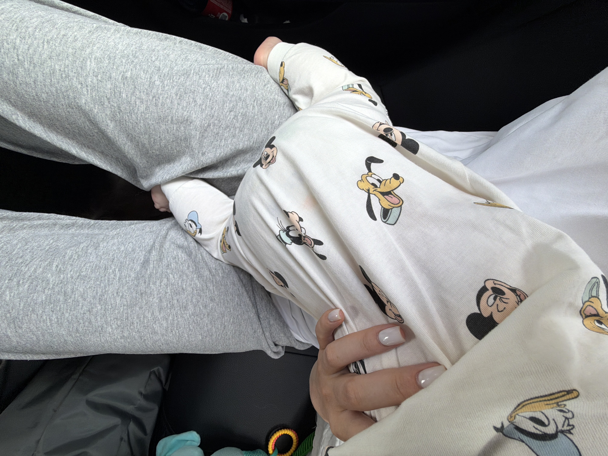 The image shows an overhead shot of a person seated, wearing gray sweatpants. A baby, dressed in white pajamas patterned with Disney characters, lies partially on the person's lap. A hand with light gray nail polish rests gently on the baby's pajamas. The scene appears to be inside a car, given the dark interior. The location is not identifiable from the image.