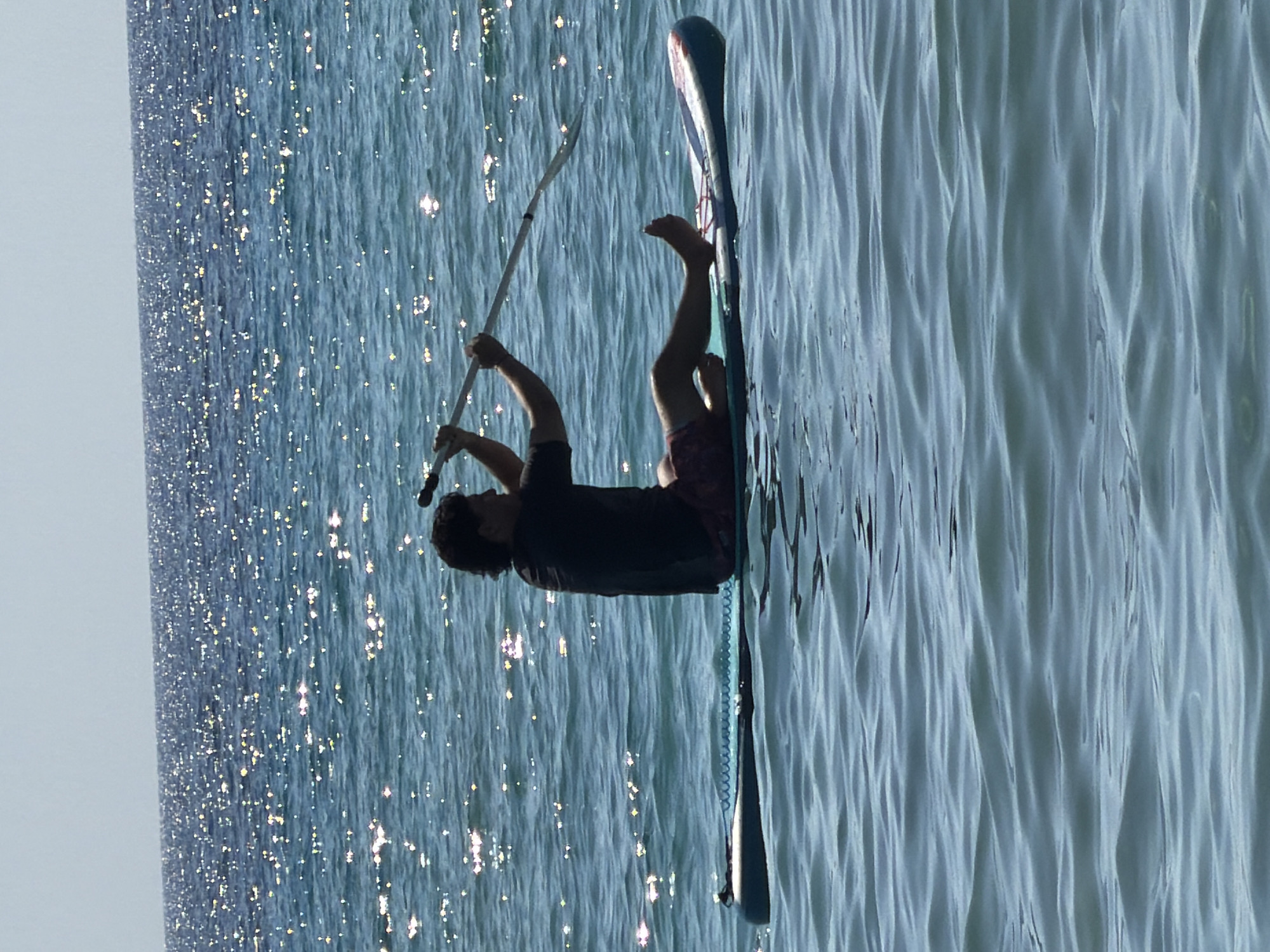 A person is seen paddleboarding on the ocean with sparkling sunlight on the water. The person is silhouetted against the bright background and is sitting on the board, holding a paddle. The water is blue and choppy, with small waves. The sky is a pale blue and clear. The scene appears to be set in a calm, sunny environment.